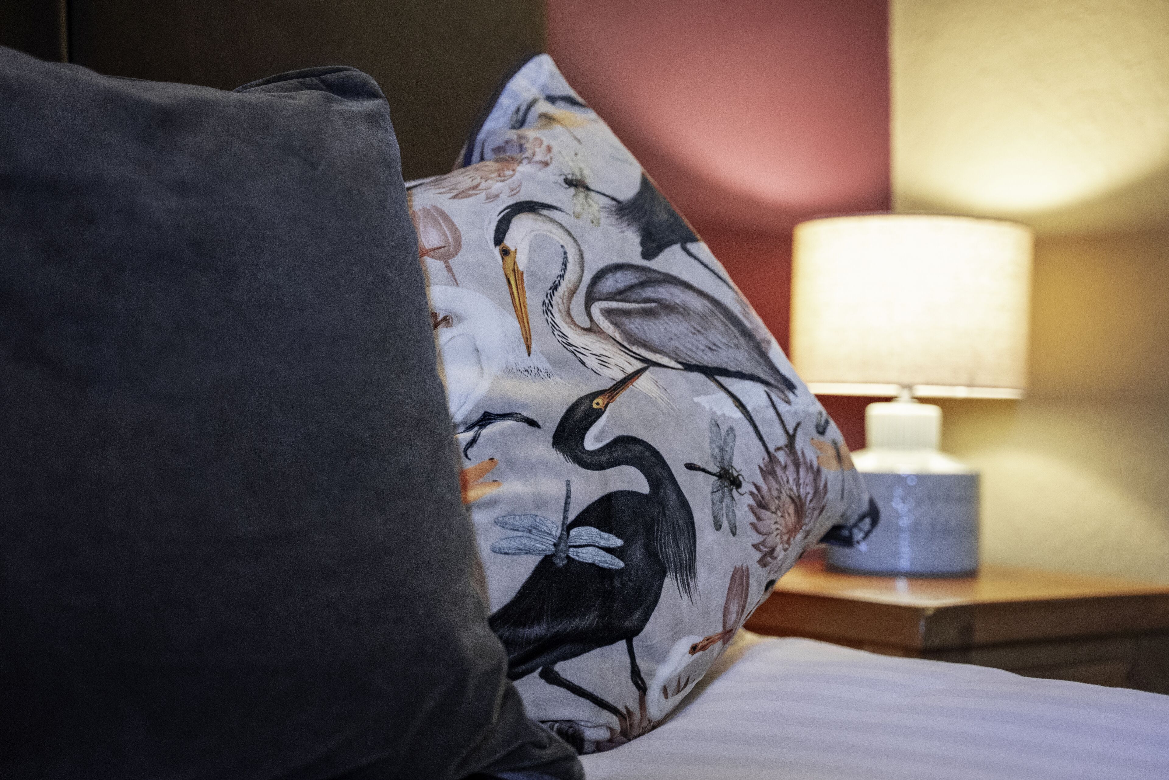 A bed with a pillow featuring a bird and floral pattern, a bedside lamp, and a wooden nightstand.
