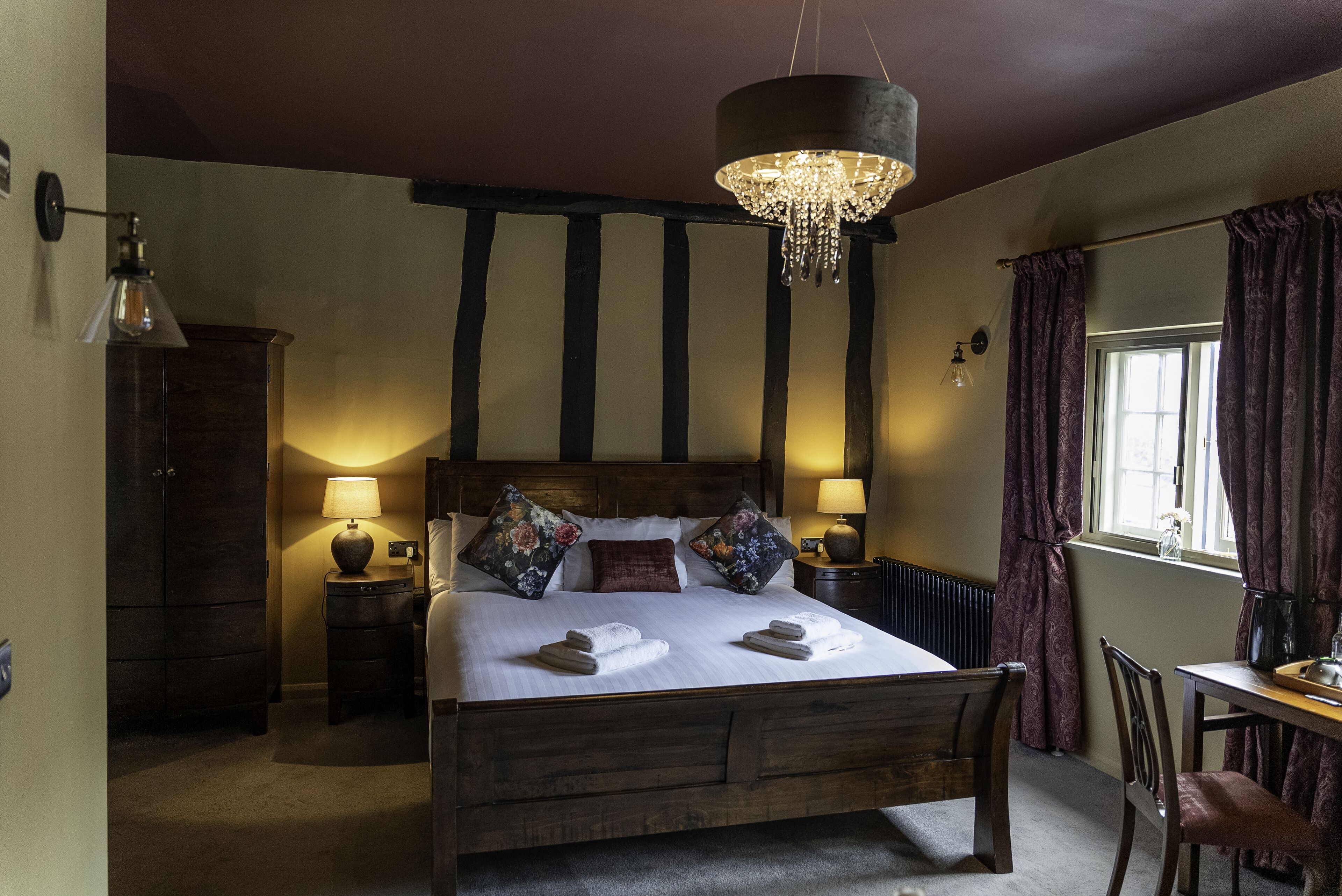 Une chambre à coucher avec un grand lit en bois, deux lampes de chevet, un lustre, une armoire en bois et une fenêtre avec des rideaux.