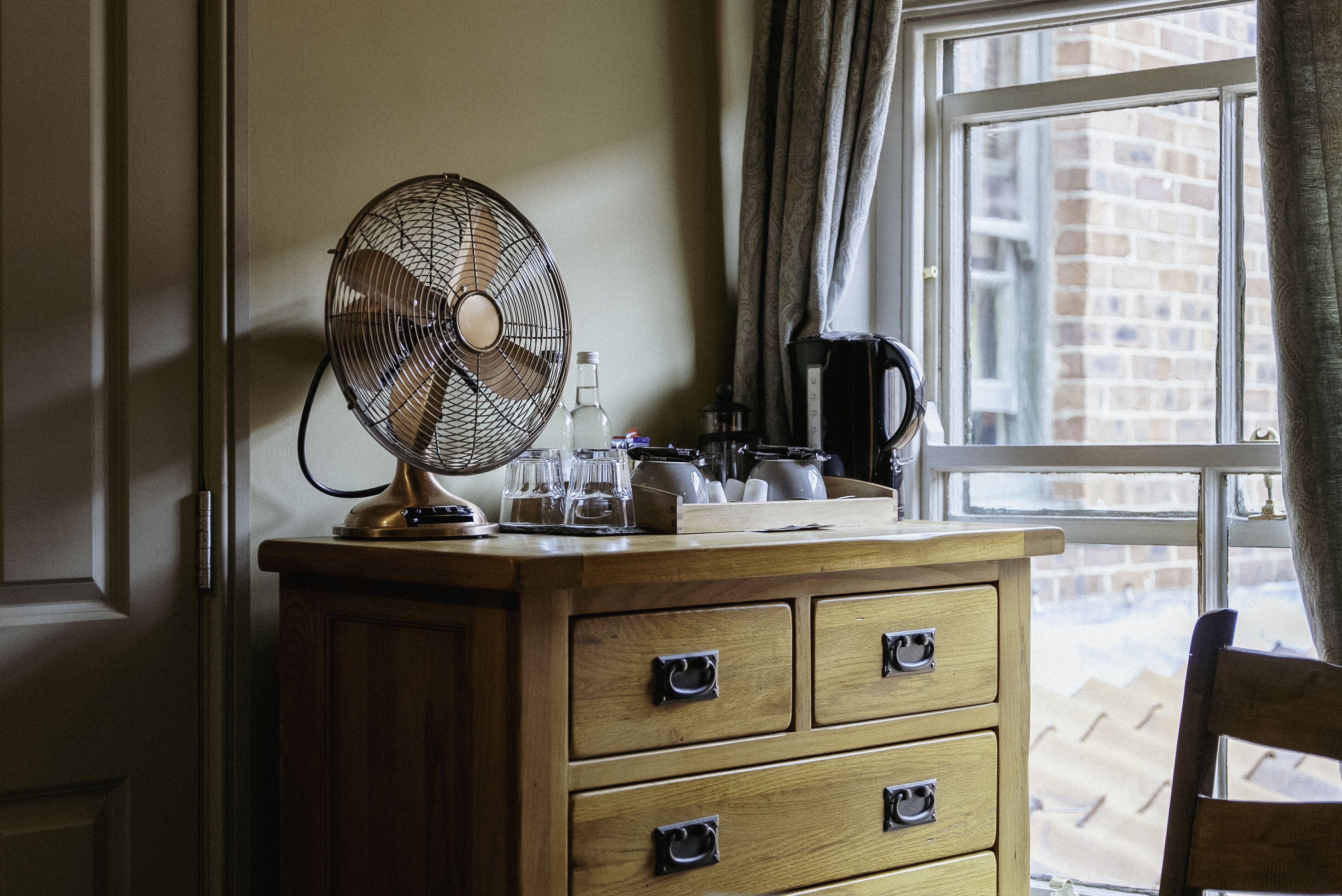 A wooden dresser with drawers, a standing fan, and a window with curtains.