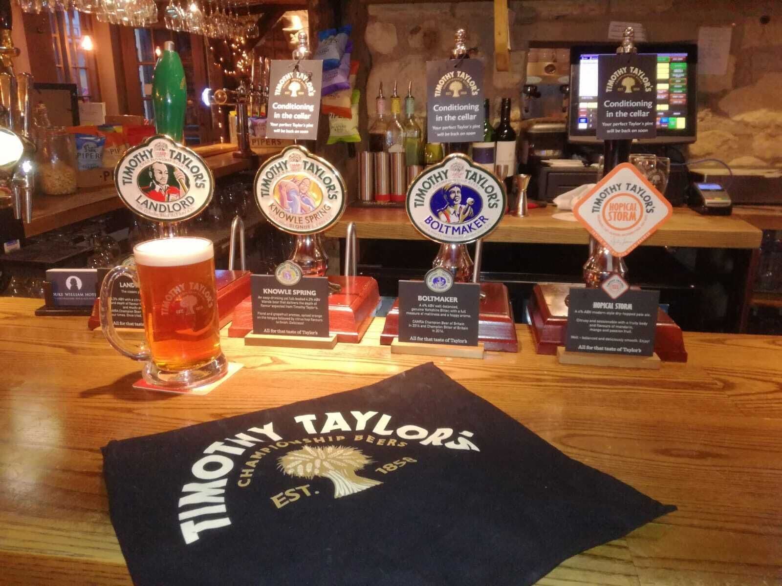 A bar area with multiple beer taps labeled Timothy Taylor's, including brands like Landlord, Knowle Spring, Boltmaker, and Tropical Storm. A glass of beer is on the counter, and a banner for Timothy Taylor Championship Beers is displayed.