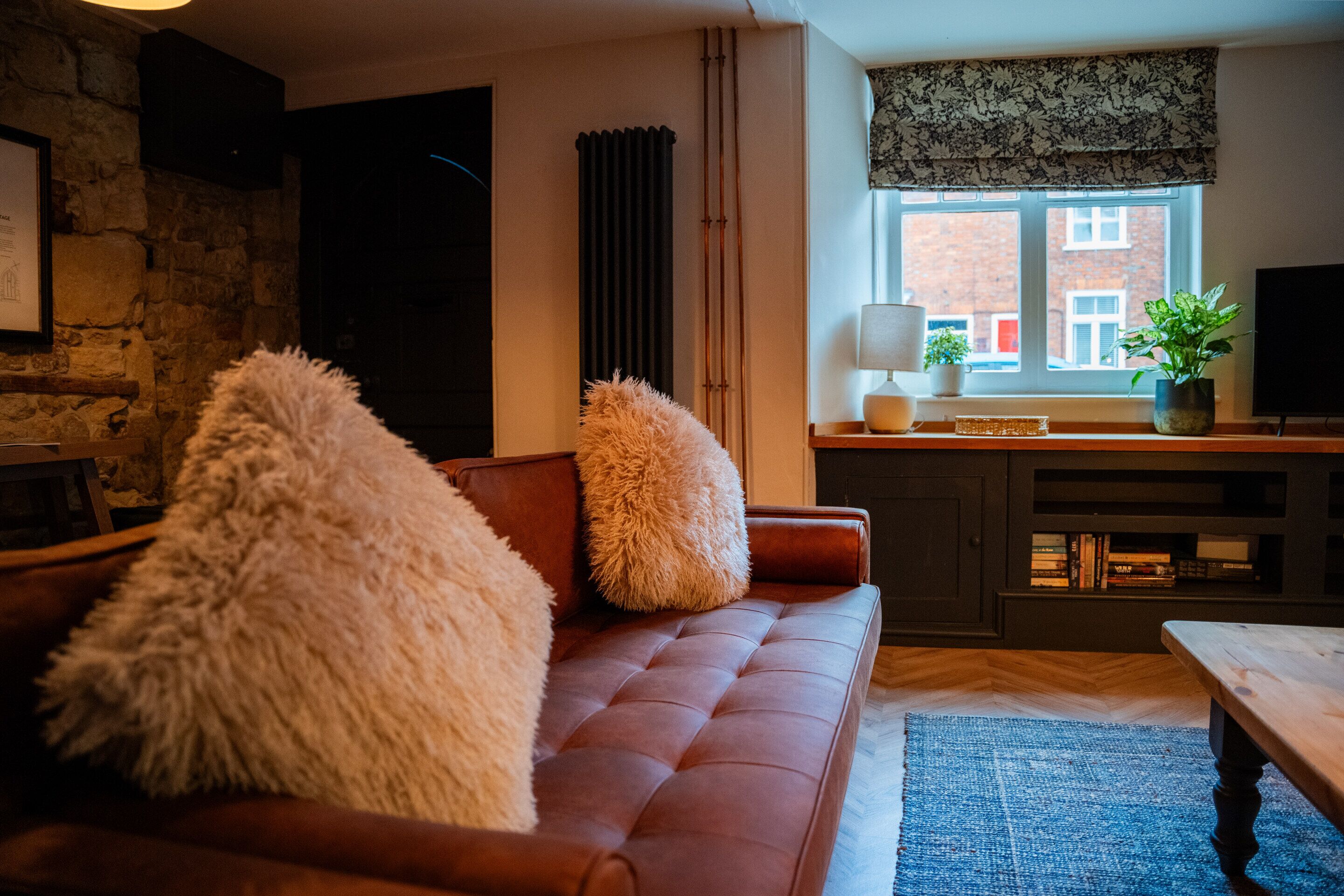 A cozy living room with a brown sofa, fluffy pillows, a blue rug, a wooden coffee table, a fireplace, a bookshelf, and a window with a view of buildings.