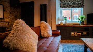 A cozy living room with a brown sofa, fluffy pillows, a blue rug, a wooden coffee table, a fireplace, a bookshelf, and a window with a view of buildings.