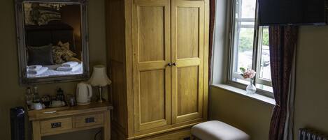 A bedroom with a wooden wardrobe, a bedside table with a lamp, a small bench, and a window with a view of the outside.
