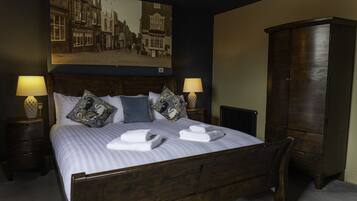 A neatly made bed with pillows and towels, a wooden headboard, a large framed picture on the wall, two bedside lamps, and a dark wooden wardrobe.