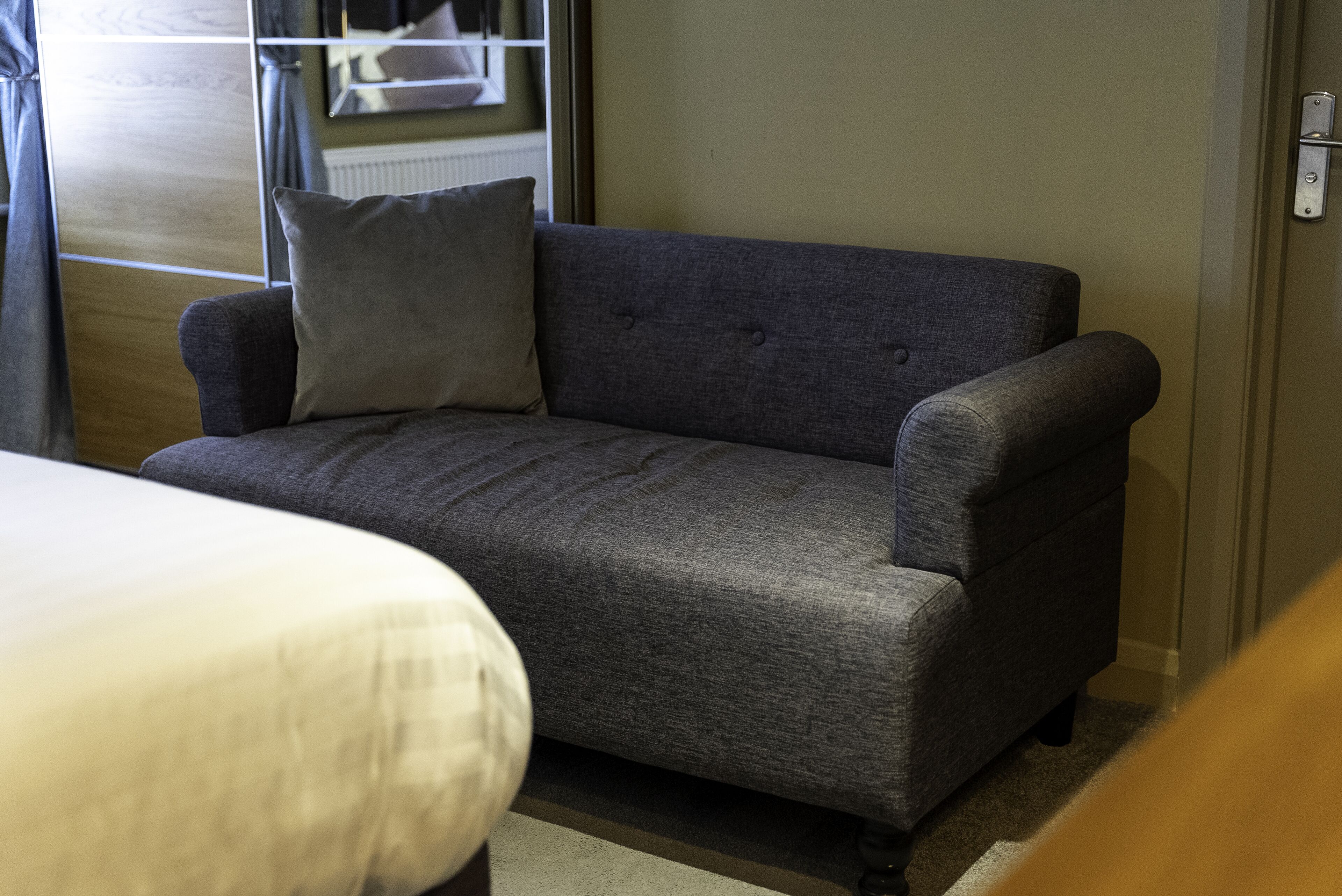 A grey sofa with a cushion, a bed with a white cover, and a window with curtains.