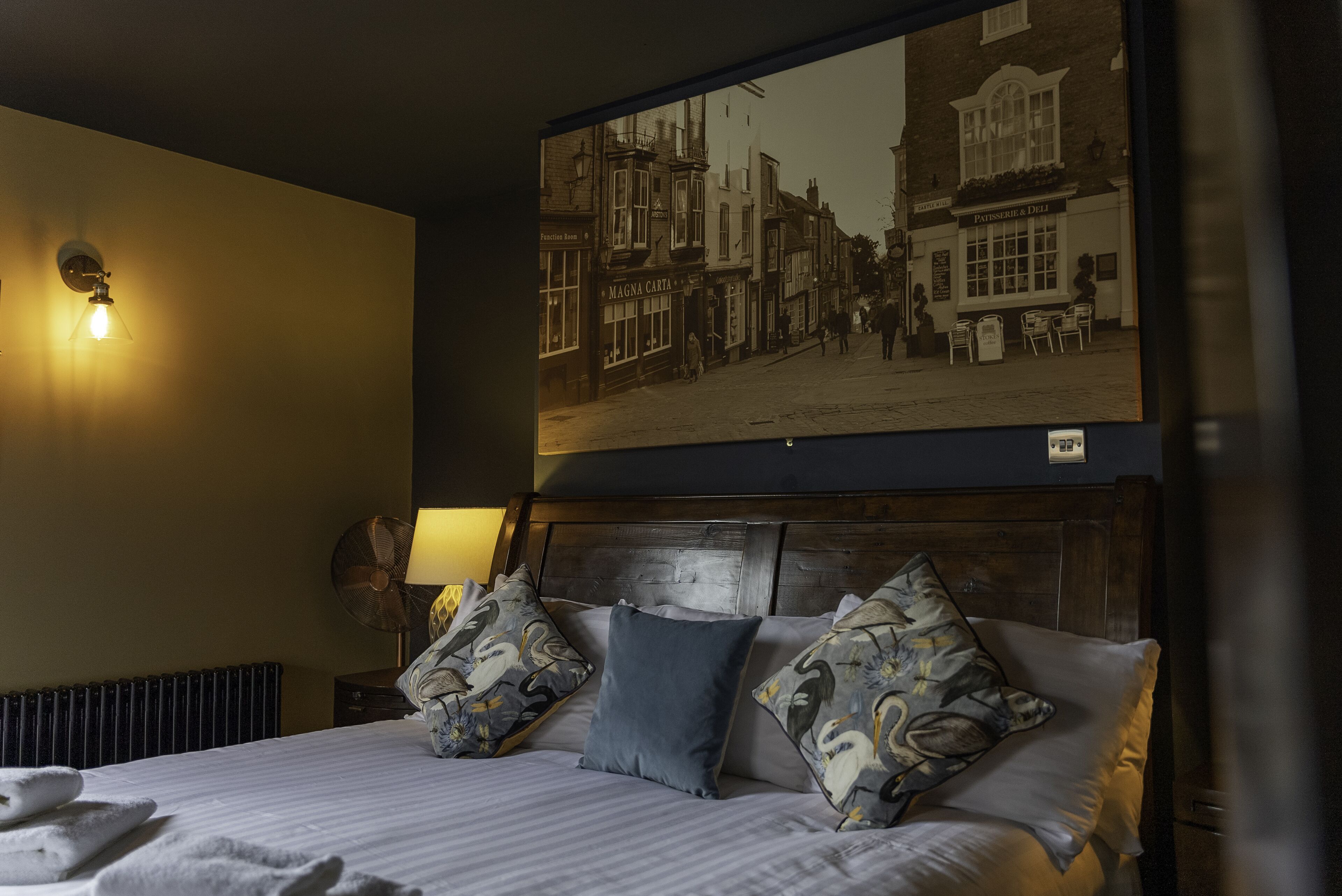 A hotel room with a bed, pillows, a wall-mounted picture, and a lamp.