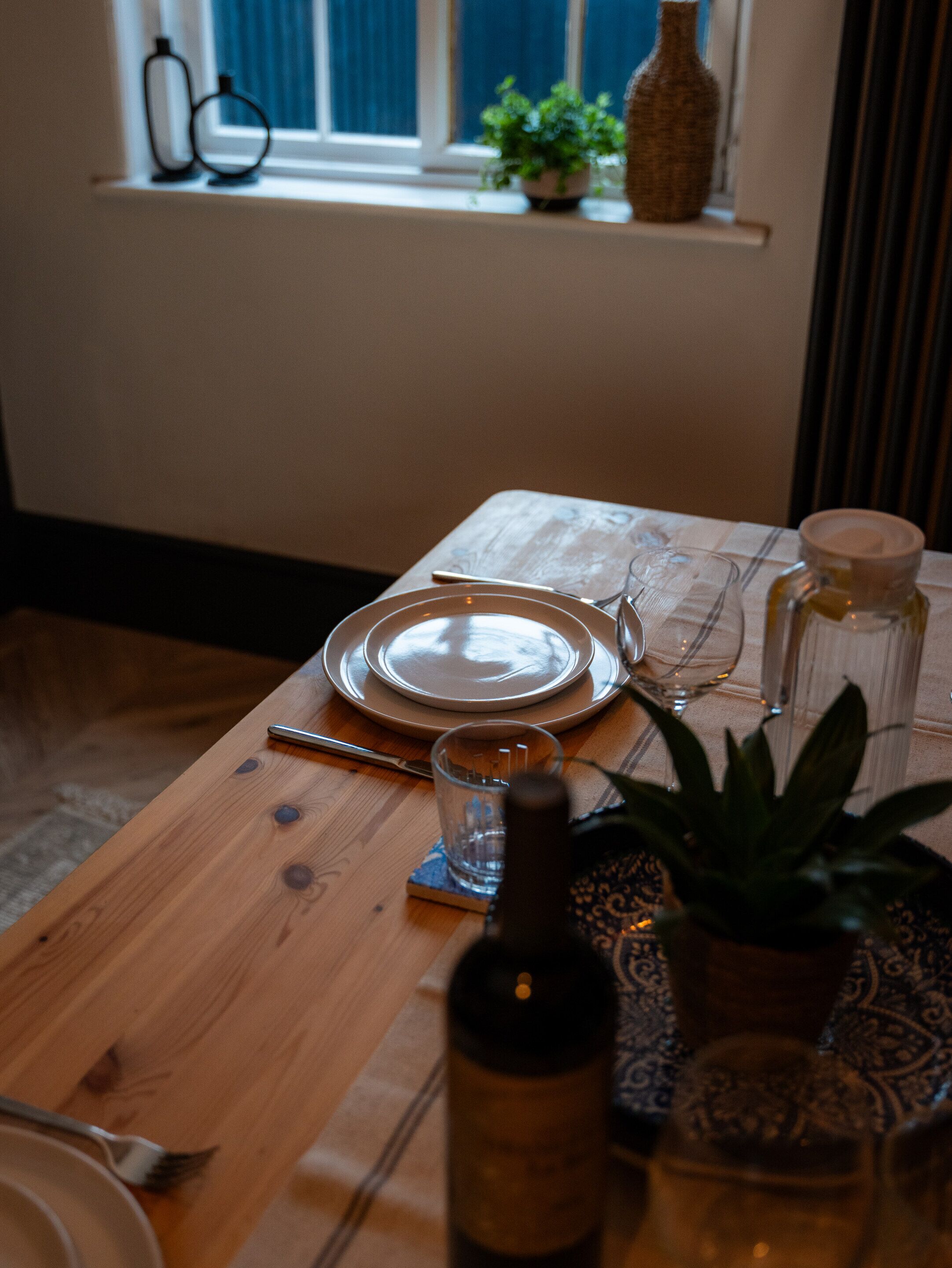 Une table à manger dressée avec des assiettes, des couverts, un verre, une bouteille et une plante en pot.