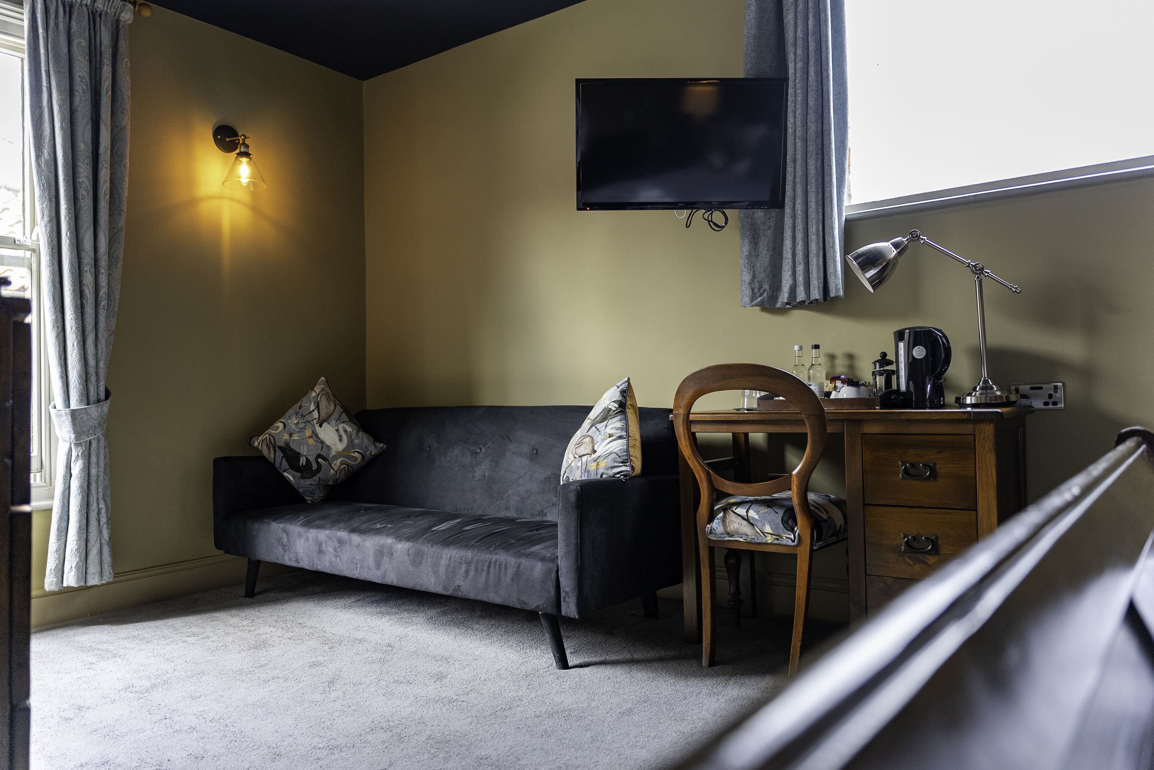 A room with a dark sofa, a wooden chair, a desk with a lamp, and a flat-screen TV mounted on the wall.