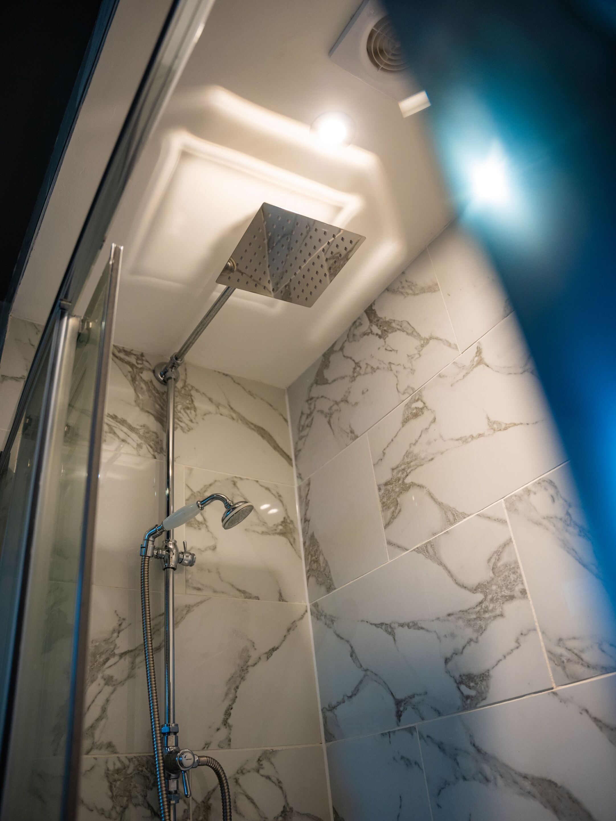 A shower with a handheld showerhead and a rain shower head, marble-patterned tiles, and a glass shower enclosure.
