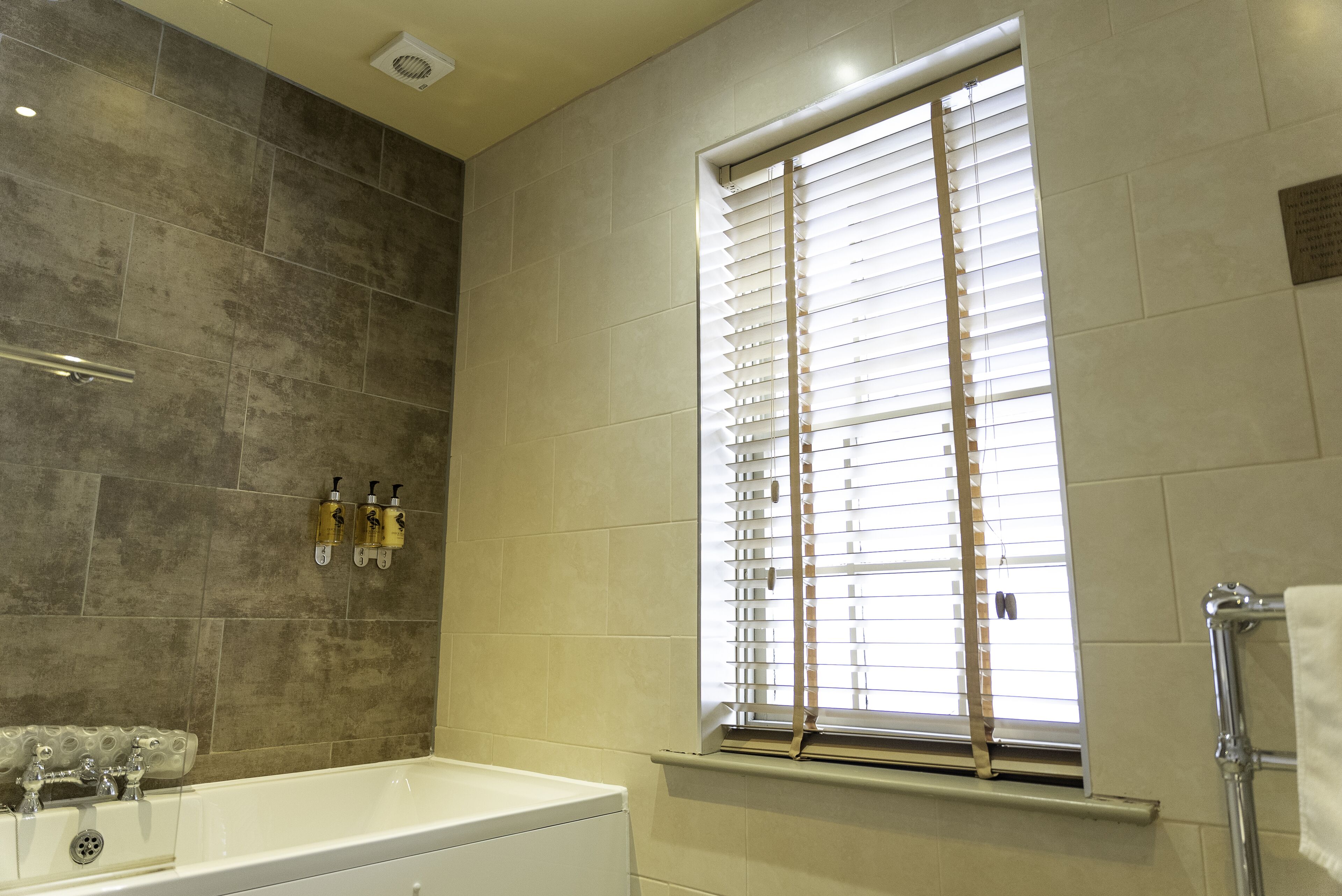 A bathroom with a bathtub, a window with blinds, and two bottles of shampoo on the wall.