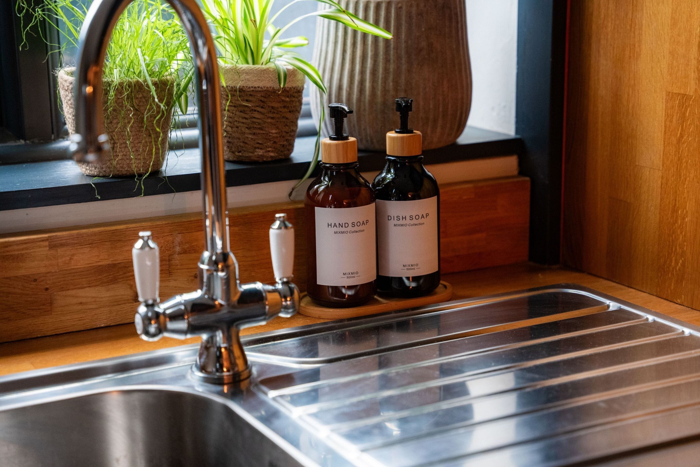 A kitchen sink with a modern faucet, two bottles of hand and dish soap, and a potted plant on the windowsill.