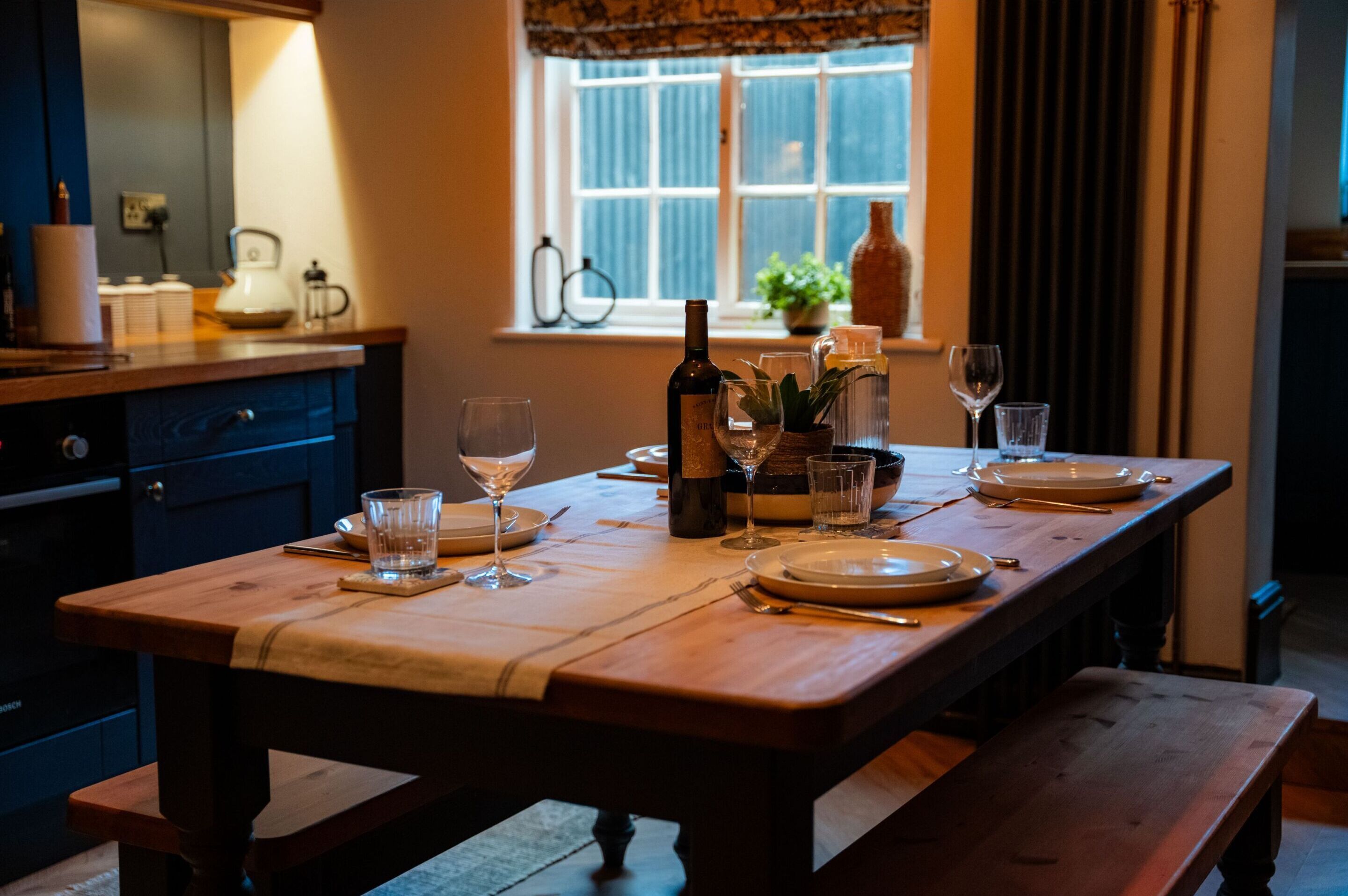 A dining table set with plates, glasses, and a bottle, with a window and curtains in the background.
