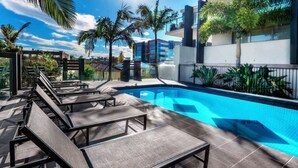 Una piscina con sillones de descanso, rodeada de palmeras y edificios modernos.