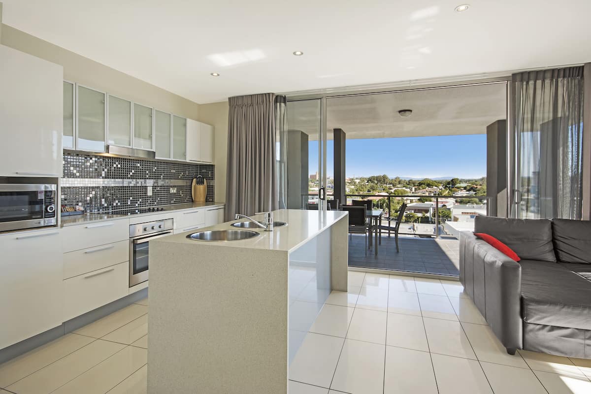 A modern kitchen with a central island, a dining area with a table and chairs, and a grey sofa with a red pillow.