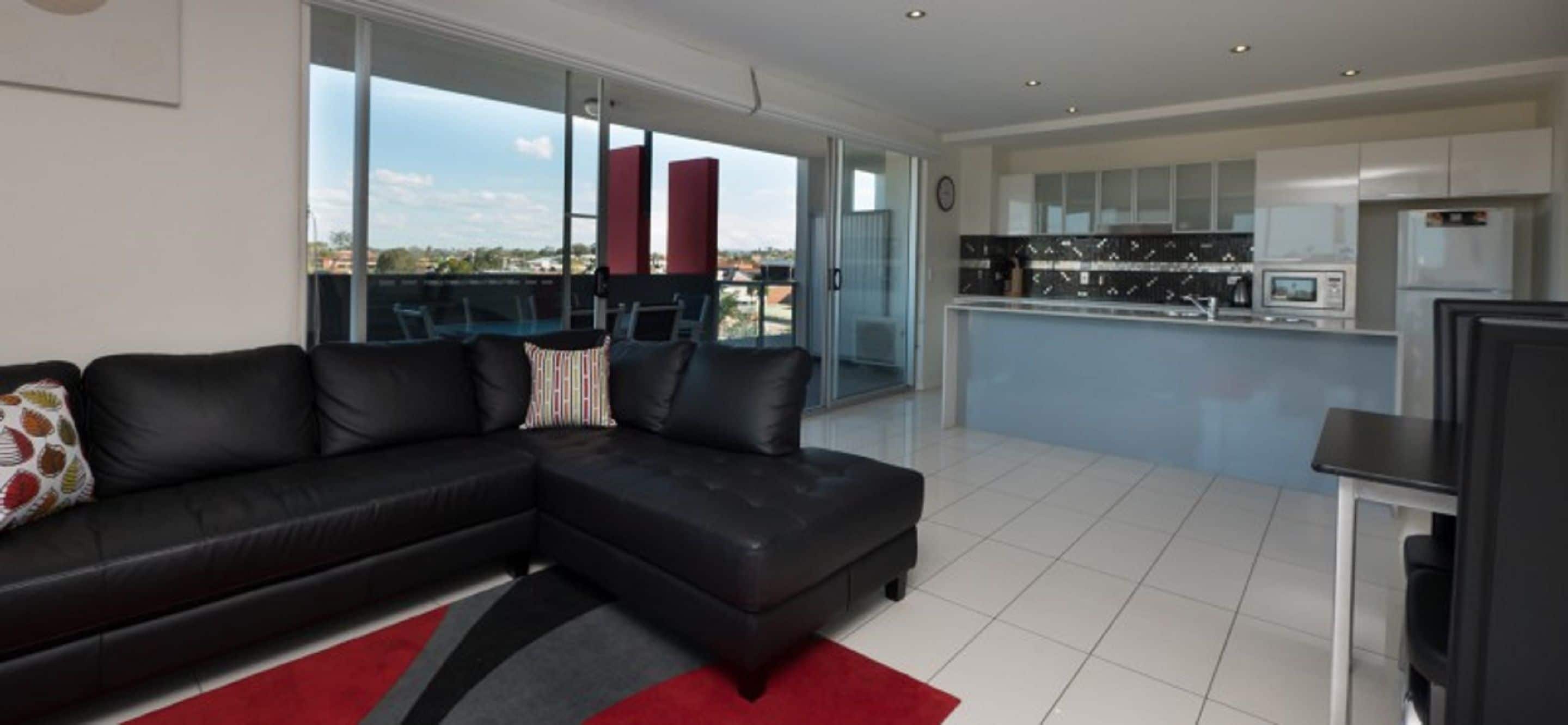 Un salon moderne avec un canapé d’angle noir, un tapis rouge et noir, et une grande porte-fenêtre coulissante donnant sur un balcon avec vue sur la ville.