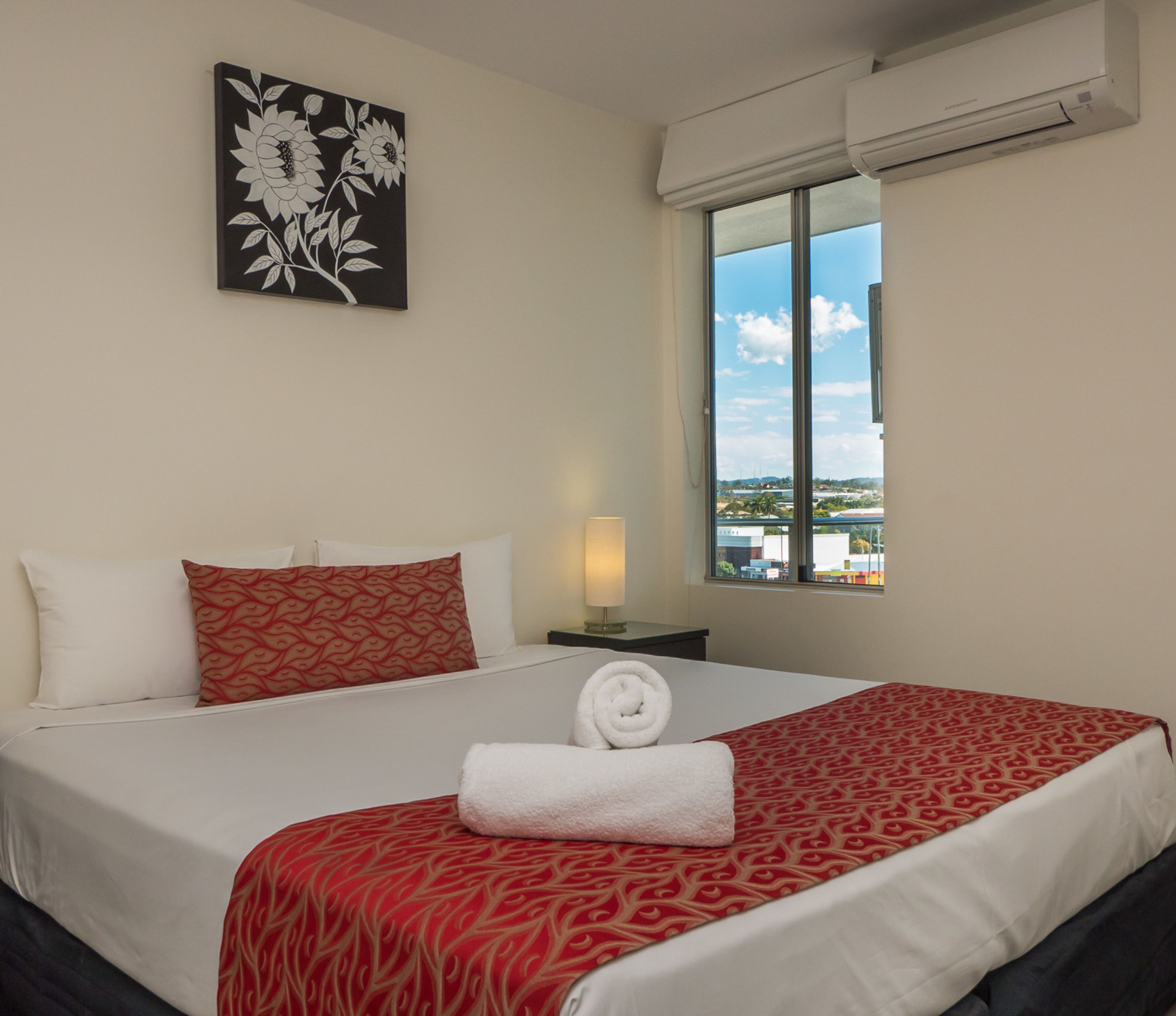 A hotel room with a bed, a window with a view, an air conditioner, and a wall-mounted artwork.