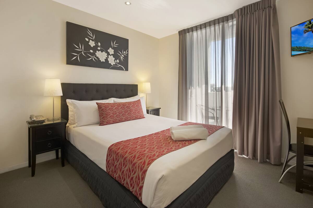 A hotel room with a large bed, two bedside tables with lamps, a flat-screen TV on the wall, and a view of the outdoors through a window with curtains.