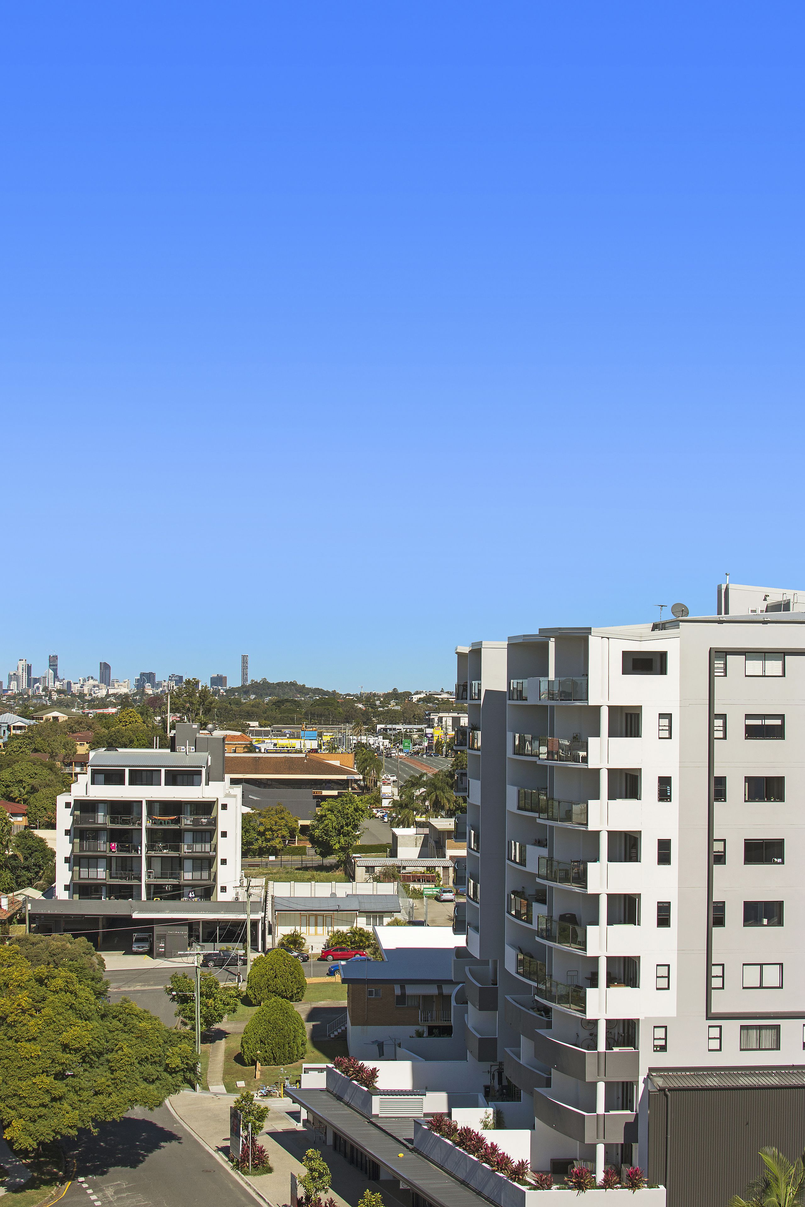 A multi-story residential building with balconies, a parking area, and a clear view of the city skyline in the background.