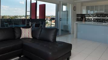 Una sala de estar moderna con un sofá modular negro, un cojín a rayas rojo y blanco, y una alfombra roja. Hay una puerta de cristal que da a un balcón con vistas a la ciudad.
