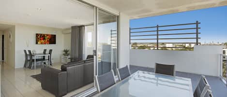 A modern living room with a glass dining table, grey sofa, and a city view through large windows.