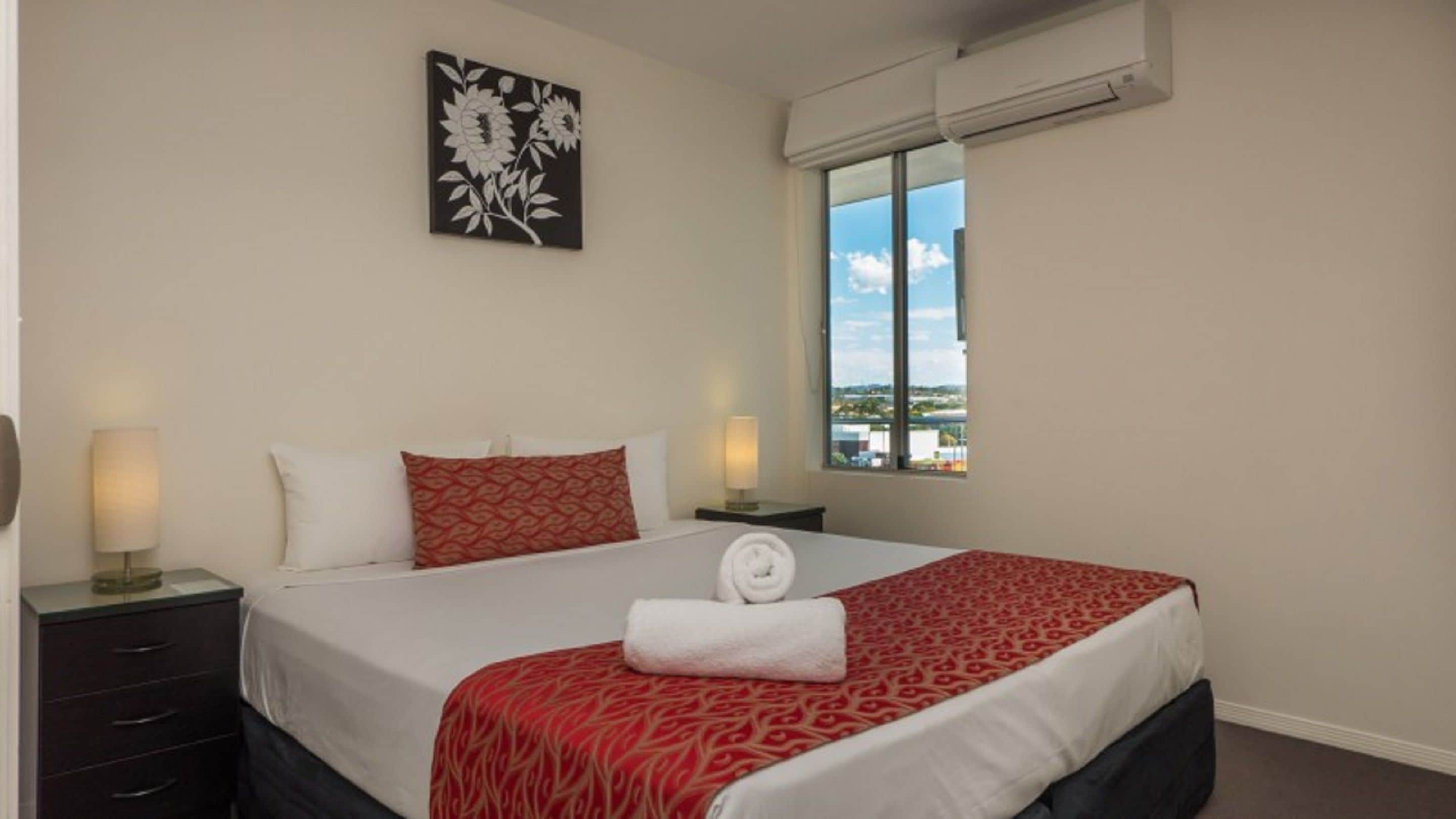A hotel room with a bed, two bedside lamps, a window with a view, and a wall-mounted air conditioner.