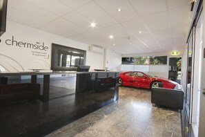 A hotel reception area with a modern design, featuring a reception desk, a red sports car, and a sign indicating 'The Chermside Boutique Hotel & Apartments'. - The Chermside Apartments (Chermside)