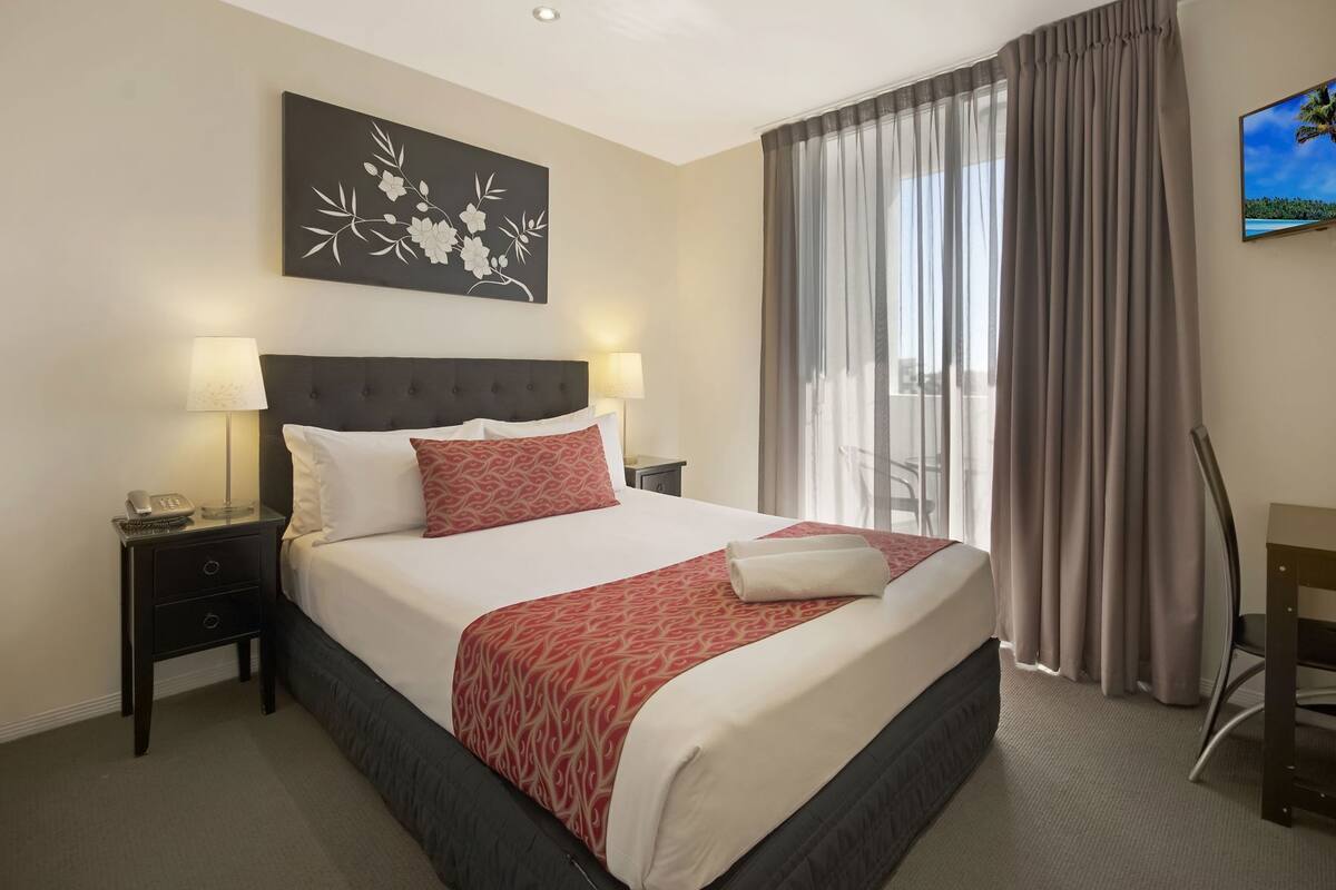 A hotel room with a bed, a nightstand, a lamp, a painting, a TV, and a view of the outdoors.
