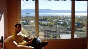 Un hombre sentado junto a un amplio ventanal con vistas a un paisaje y al agua.