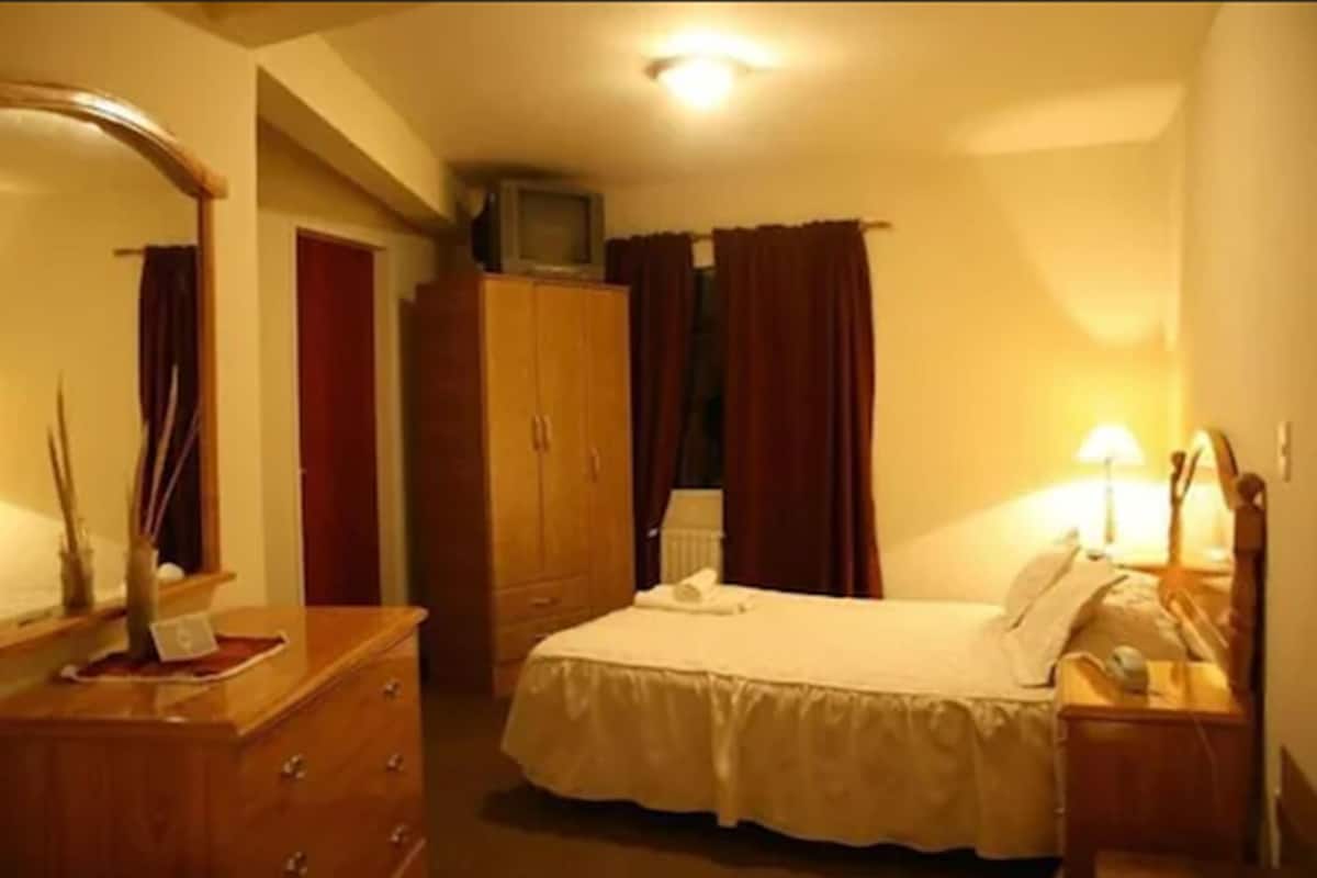 Une chambre d’hôtel avec un lit, une armoire en bois, un téléviseur posé sur un meuble et une table de chevet avec une lampe.