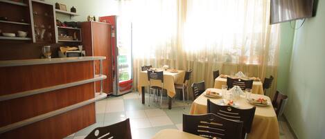 Zona pranzo con tavoli apparecchiati per quattro persone, una macchina distributrice e scaffali con stoviglie.