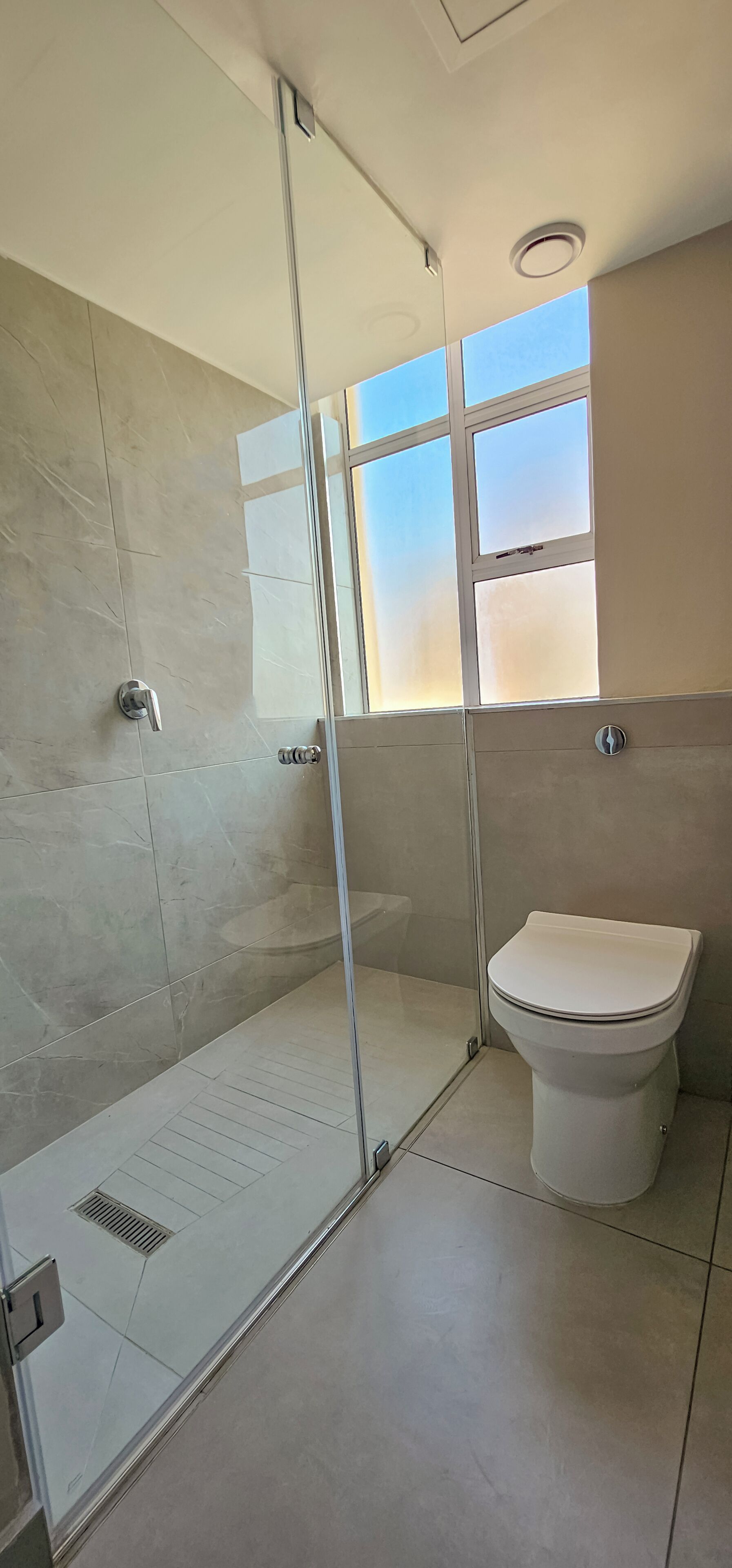 A modern bathroom with a glass shower enclosure, a toilet, and a window providing natural light.