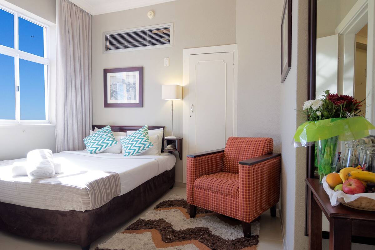 Een hotelkamer met een bed, een stoel, een bureau met een bloevas en een raam met gordijnen.