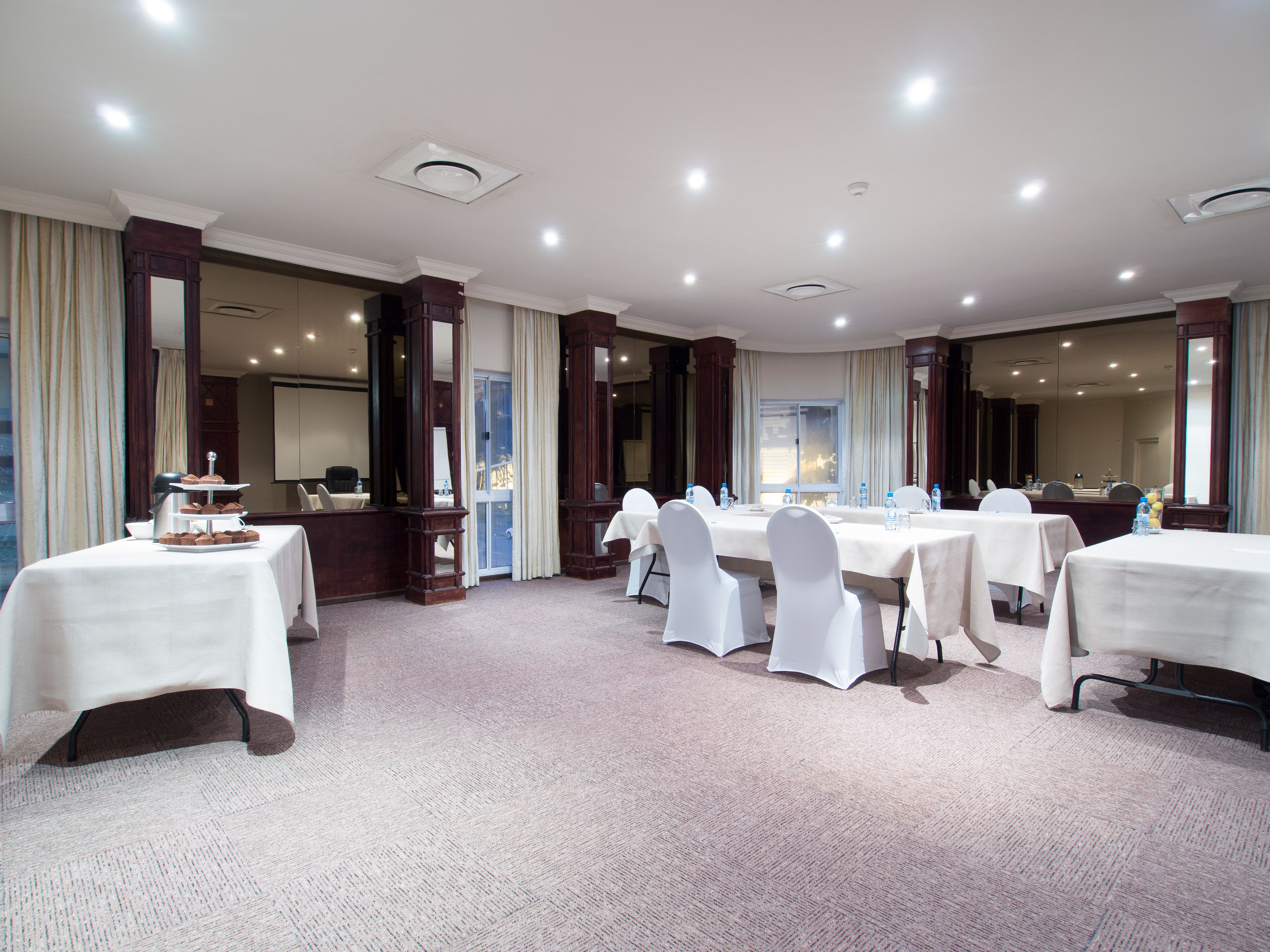A conference room with multiple tables and chairs, a buffet table with pastries, and a large window with curtains.