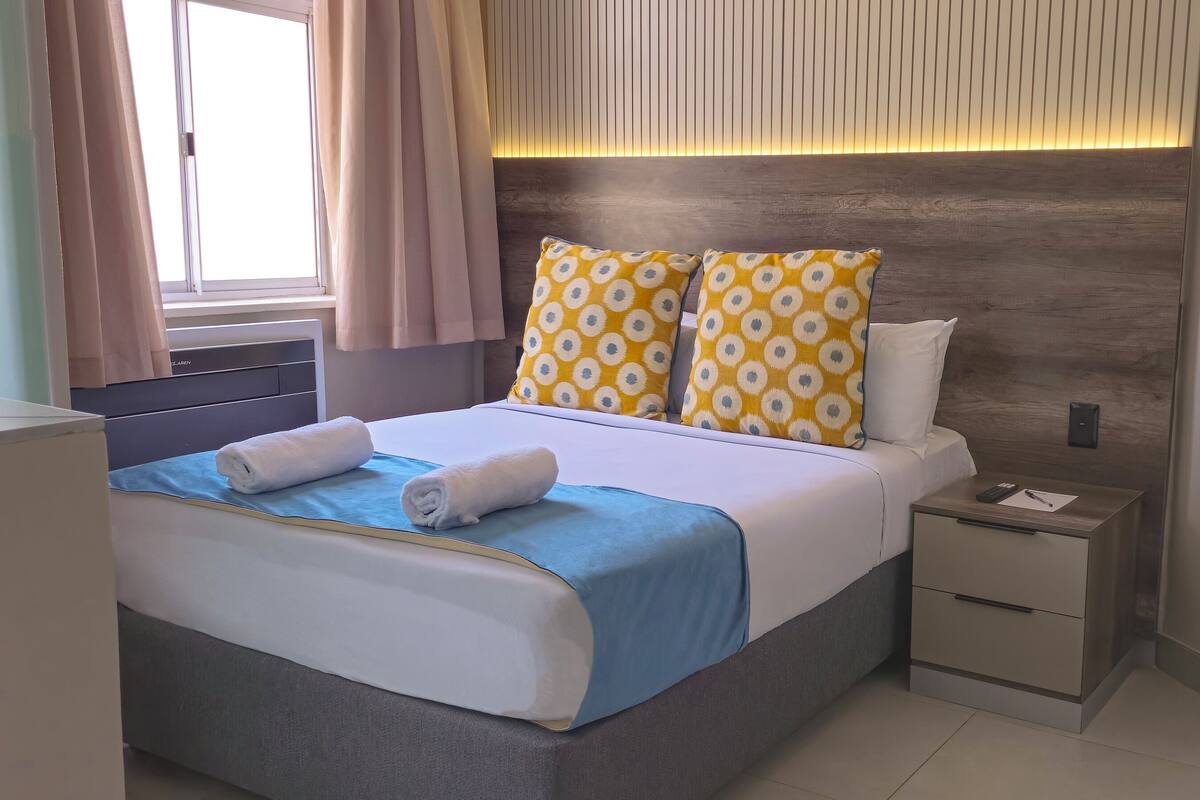 Een netjes opgemaakt bed met een blauw-witte dekbedovertrek, twee kussens met een geel-wit patroon, en een nachtkastje met een afstandsbediening en papieren.
