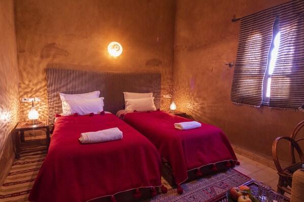 Une chambre avec deux lits, chacun recouvert d’une couverture rouge et agrémenté d’oreillers blancs, une petite table avec une lampe, et une fenêtre ornée de rideaux à rayures.