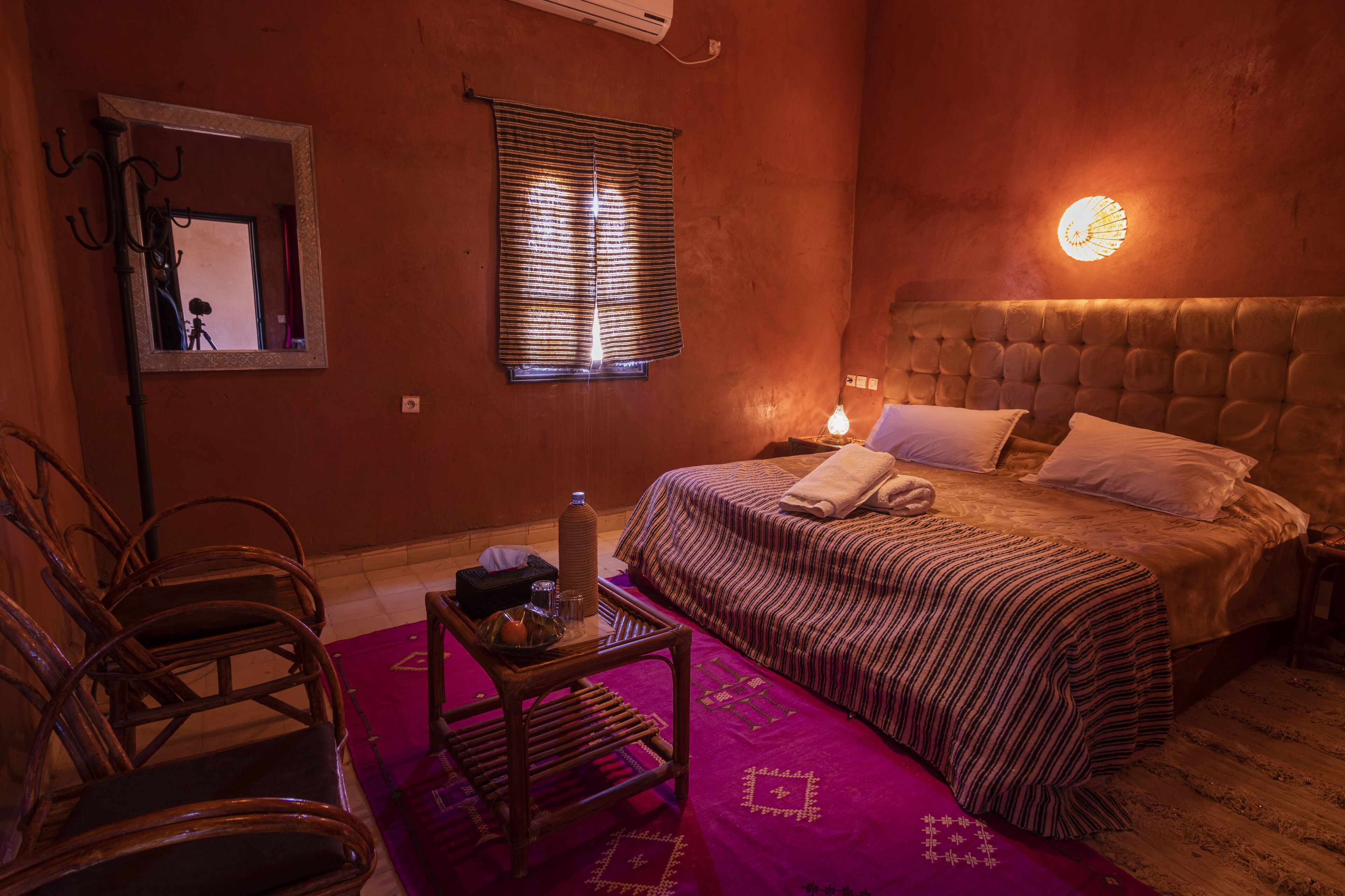 A bedroom with a bed, a chair, a table, a window with blinds, and a wall-mounted light.