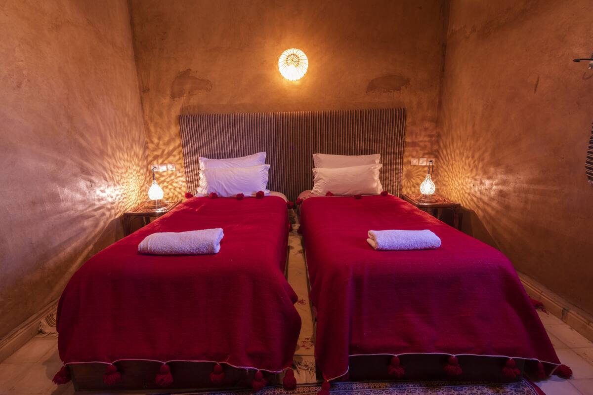 Une chambre avec deux lits, chacun recouvert d’une couverture rouge et orné d’oreillers blancs, et un seul luminaire au plafond.