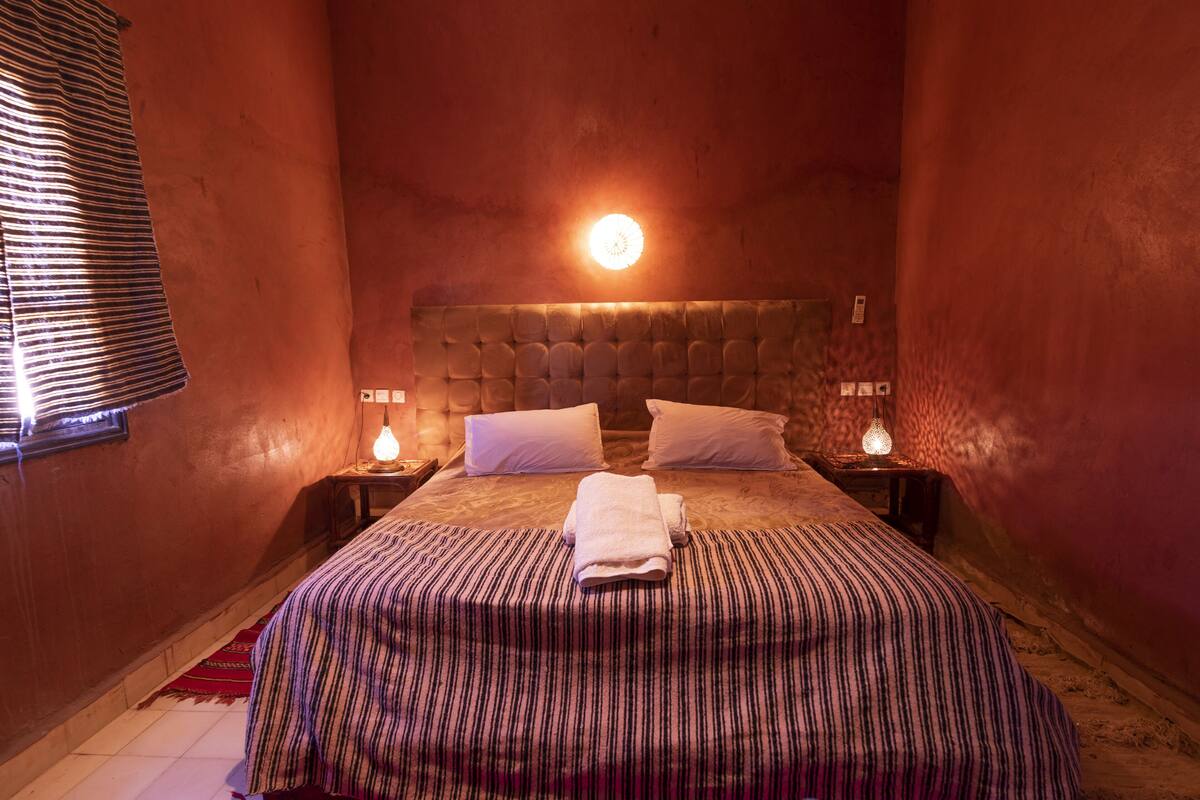 Une chambre à coucher accueillante, avec une tête de lit capitonnée, une couvre-lit à rayures, deux oreillers blancs et une lampe de chevet.