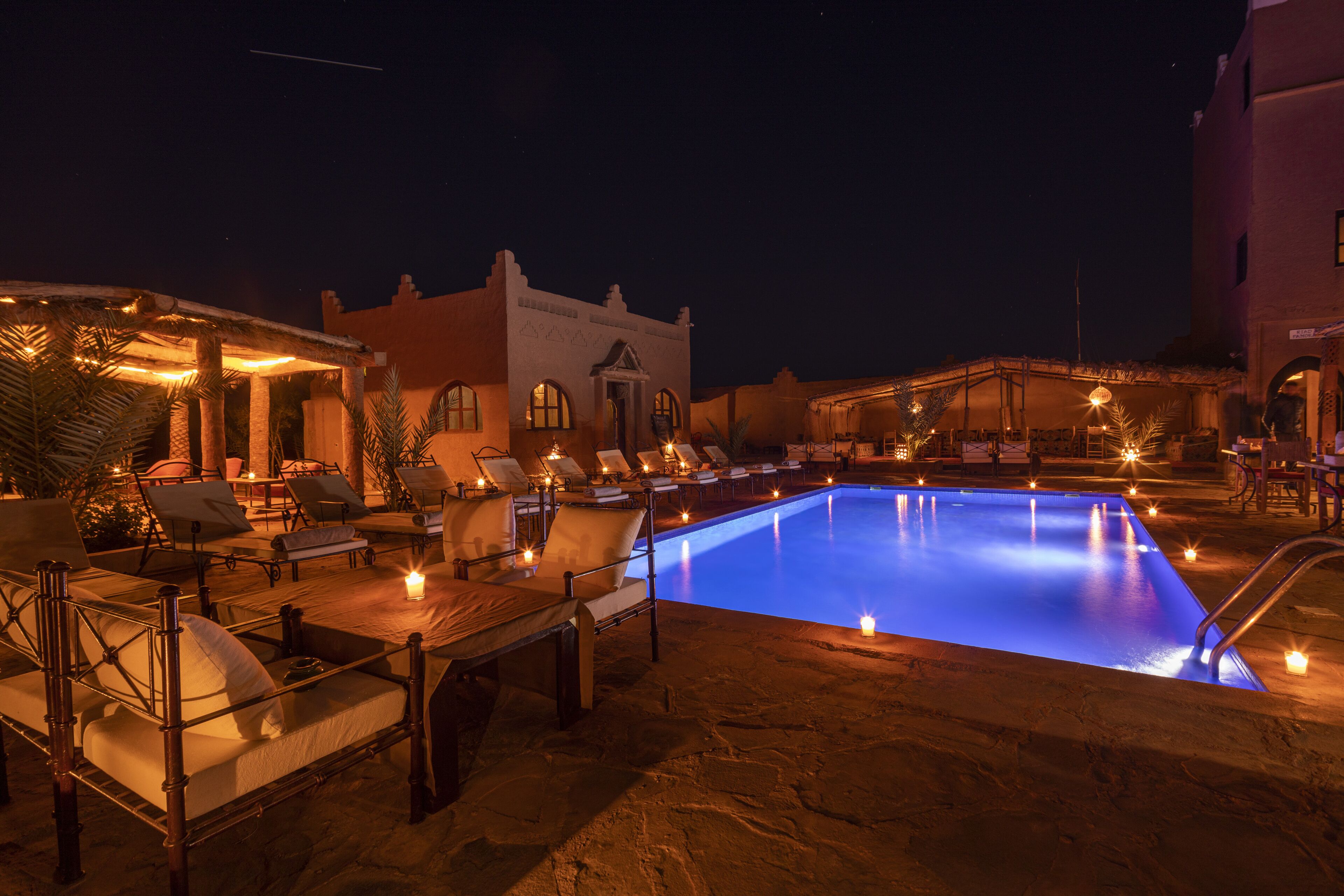 A nighttime pool area with lounge chairs and tables, a building with arched windows, and a clear pool.