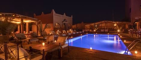 A nighttime pool area with lounge chairs and tables, a building with arched windows, and a clear pool.