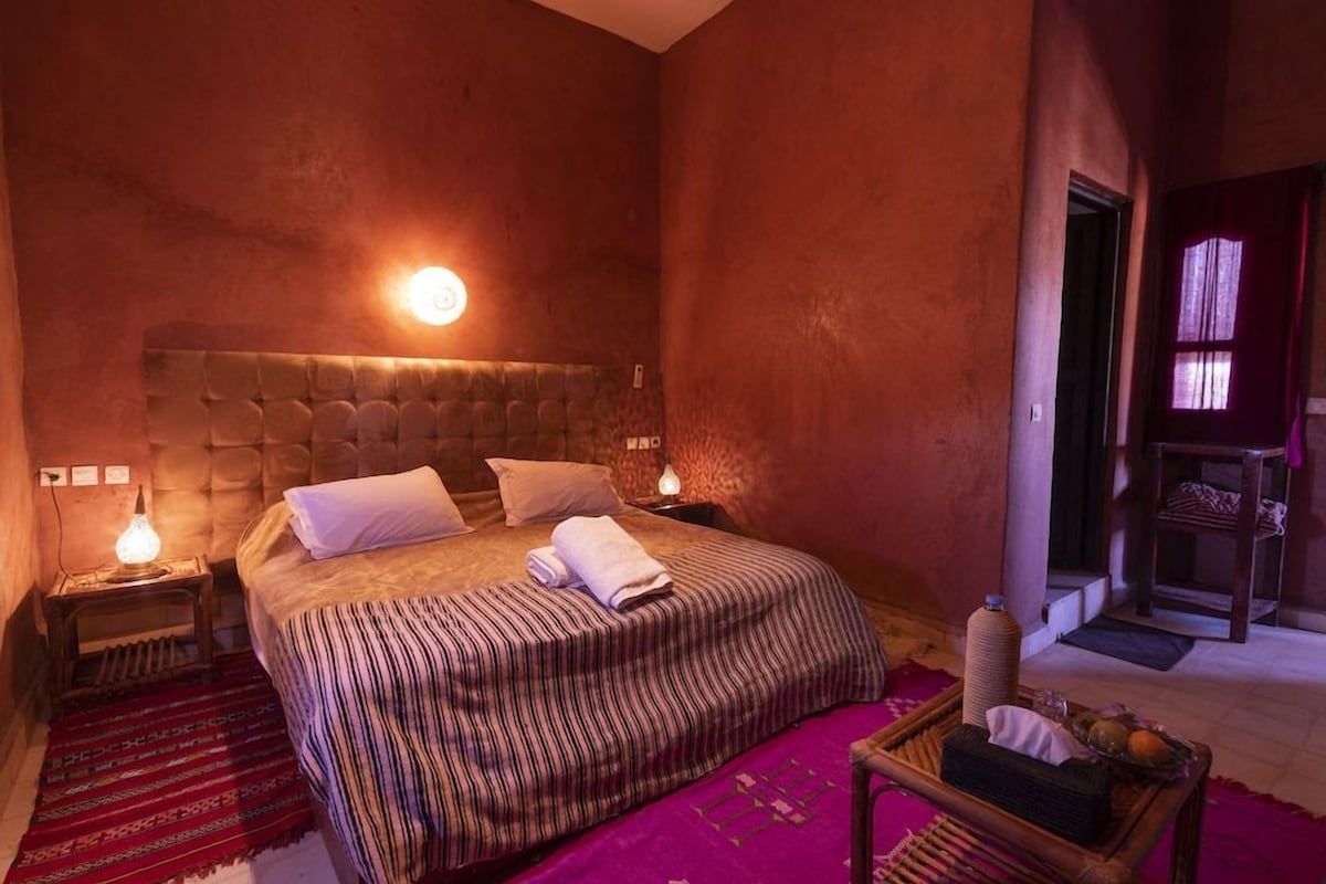 A bedroom with a large bed, two bedside lamps, a nightstand, a red patterned rug, and a small table with a basket.