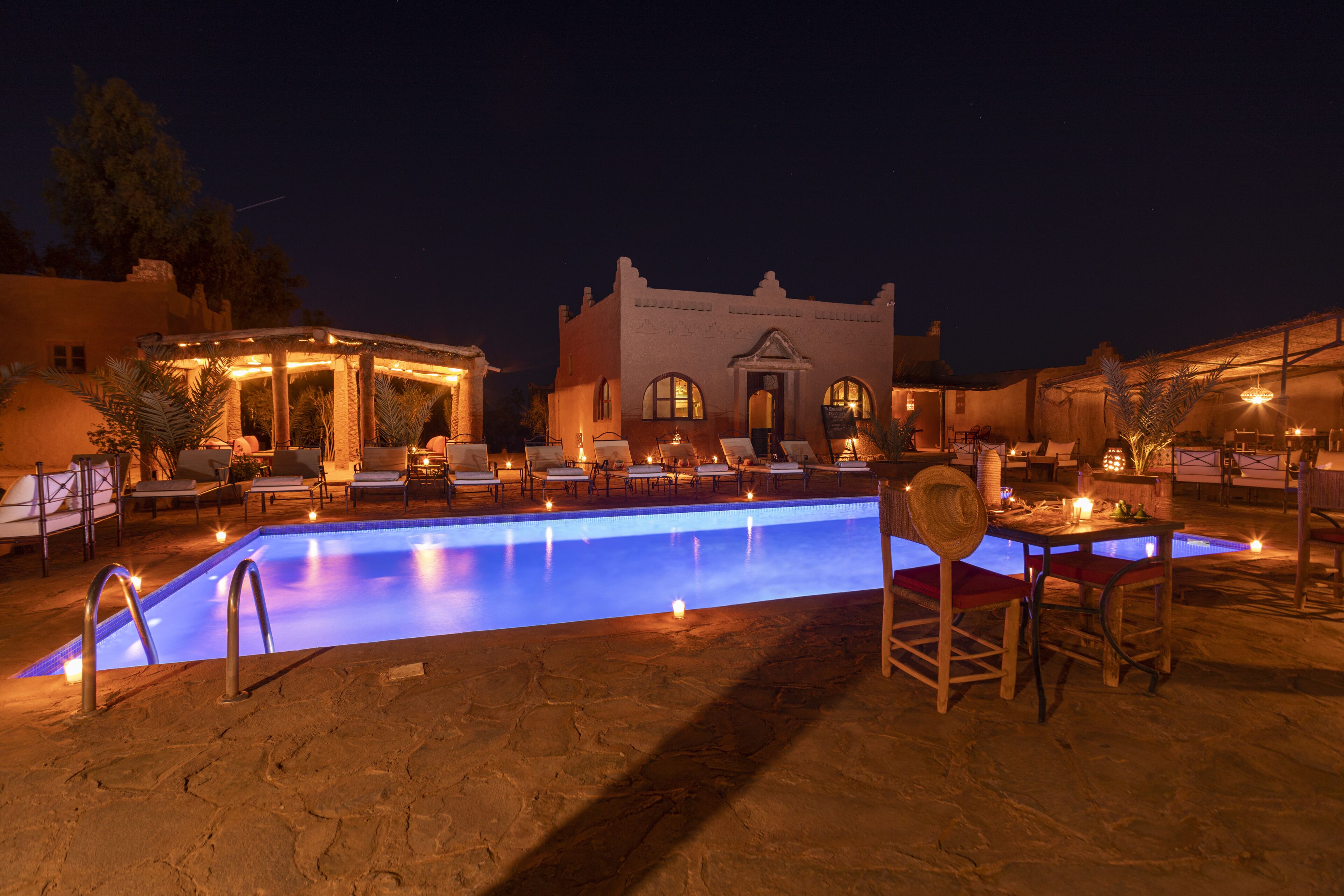 Un espace piscine avec des chaises longues et des tables, un bâtiment doté de fenêtres en arc de cercle et un ciel nocturne dégagé.