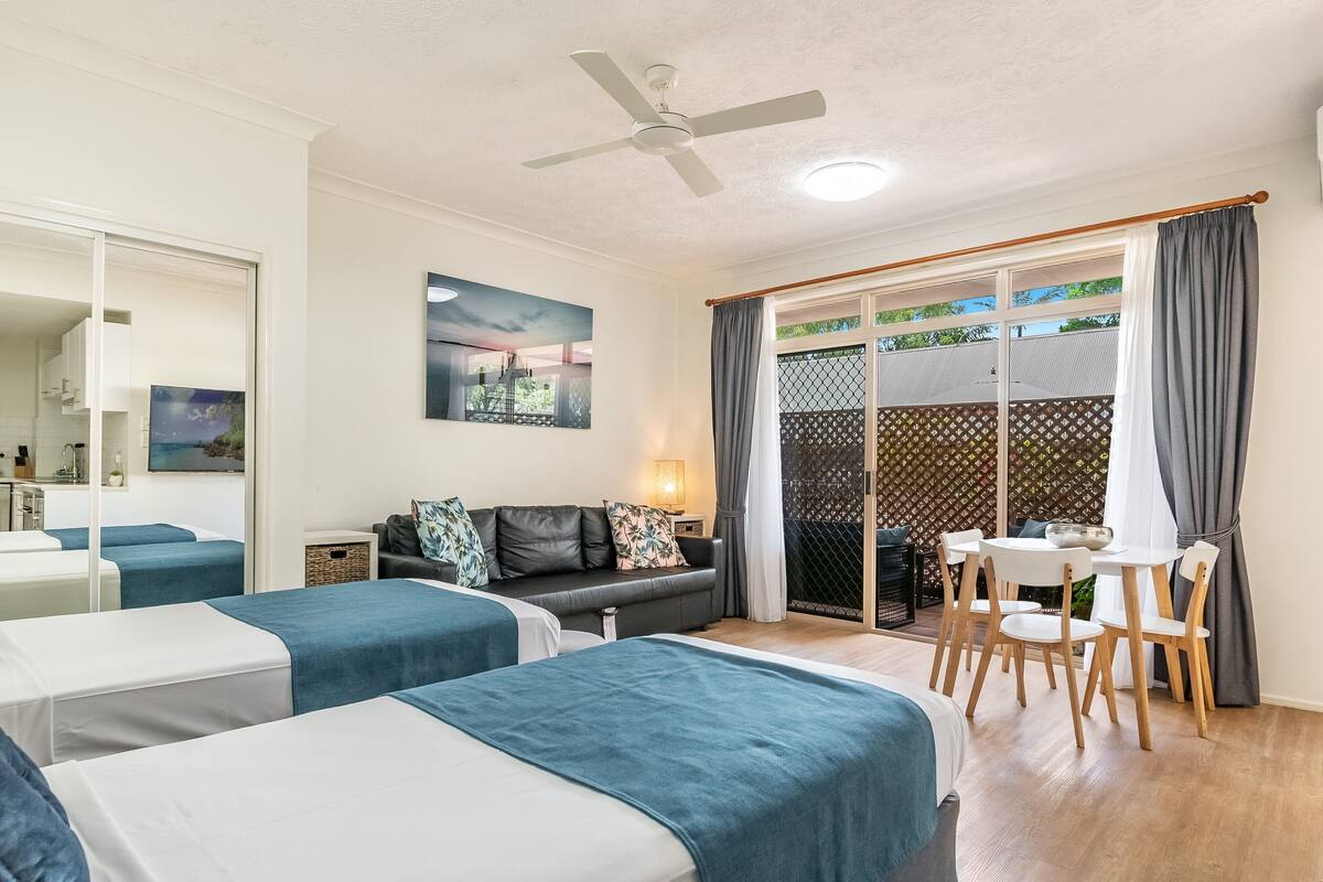 A hotel room with two beds, a sofa, a dining table with chairs, a ceiling fan, and a view through a sliding glass door.