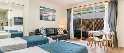 Habitación de hotel con dos camas, un sofá, una mesa de comedor con sillas, un ventilador de techo y vistas a través de una puerta corrediza de cristal.