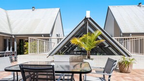 A patio area with a glass table and metal chairs, surrounded by potted plants and a view of two buildings with triangular roofs. - Byron Central Apartments (Byron Bay)