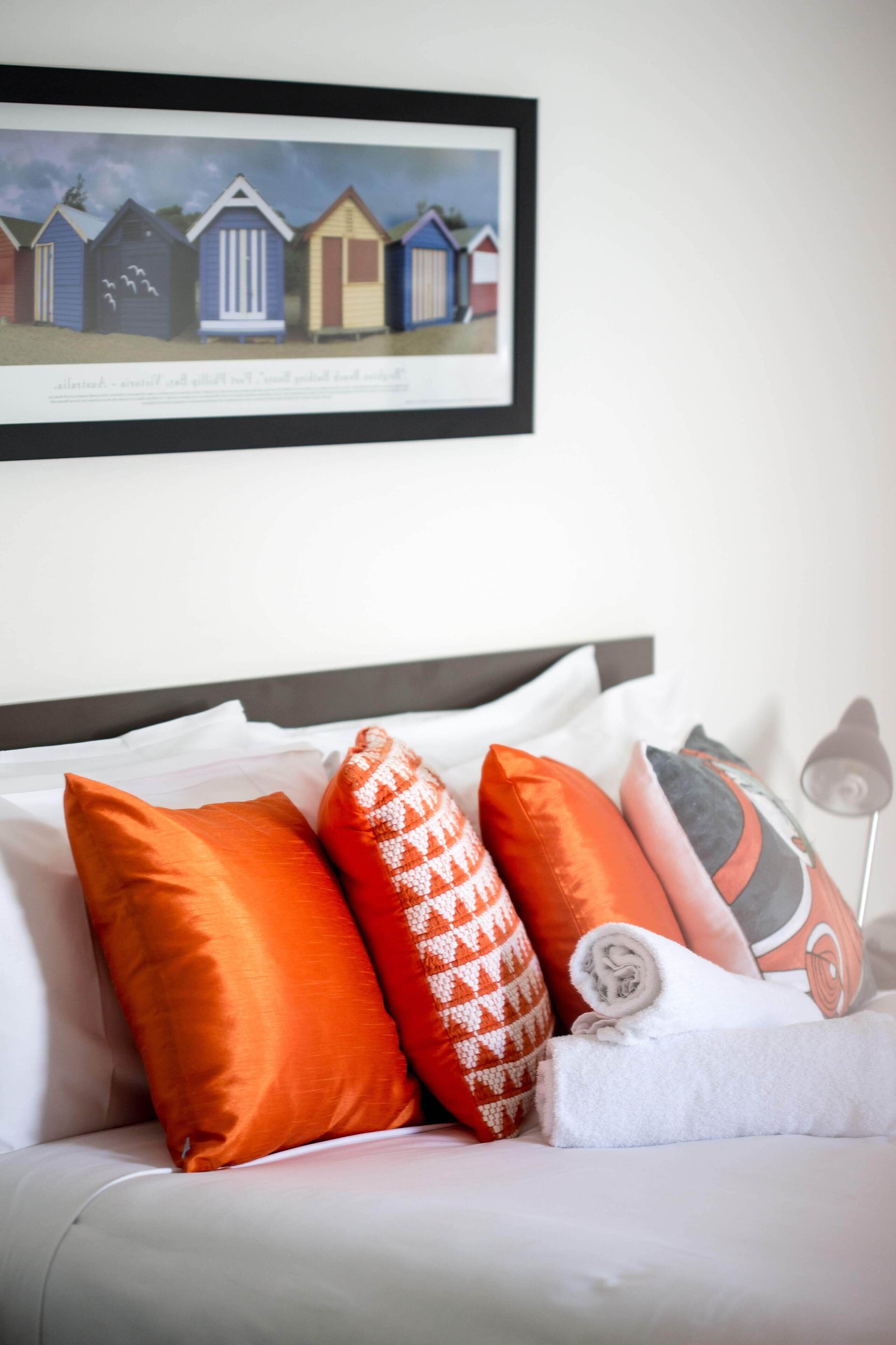 A bed with white bedding and orange pillows, a framed picture of beach huts on the wall, and a lamp on the side.