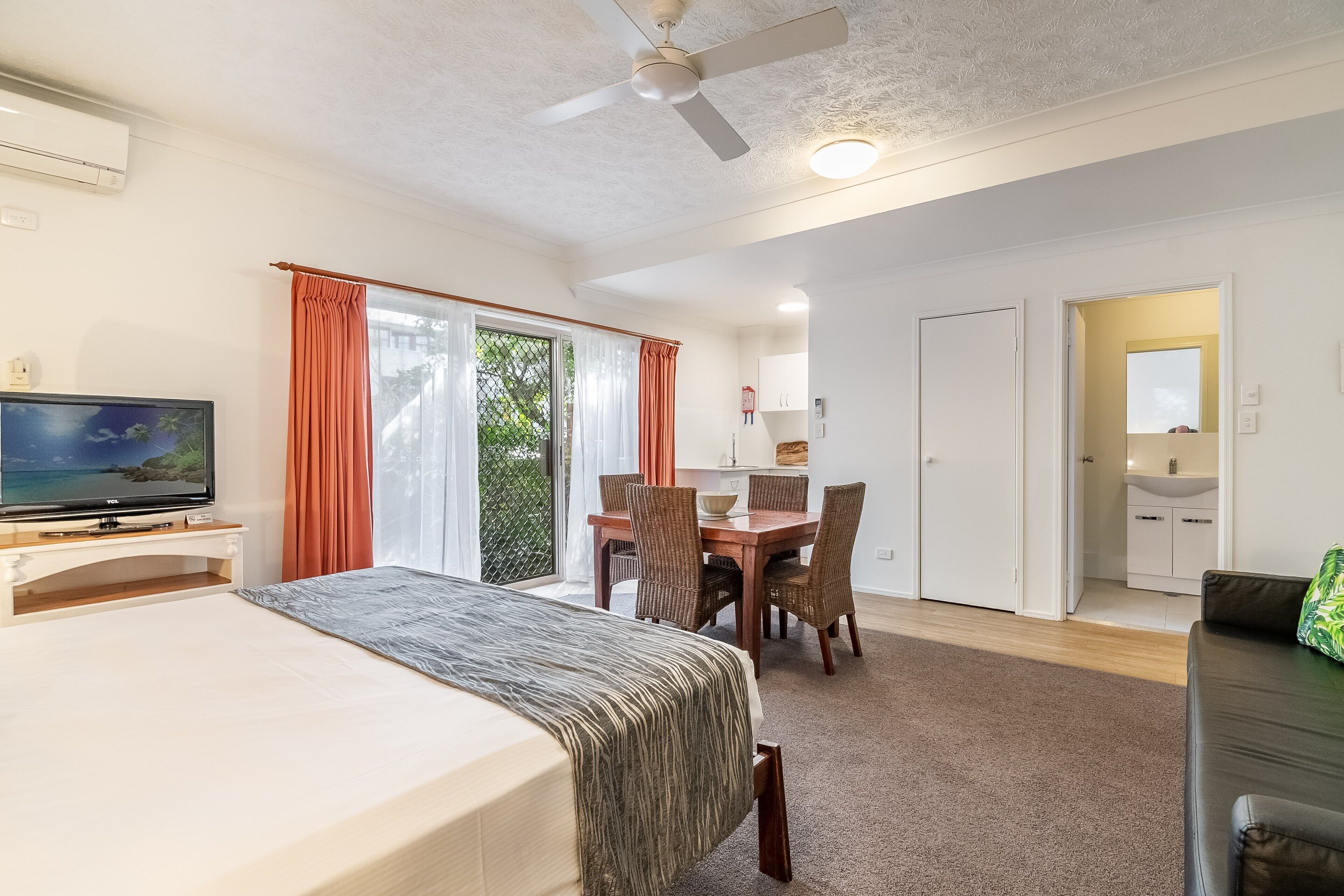 Habitación de hotel con cama, televisor, mesa de comedor, sillas y balcón con cortinas.