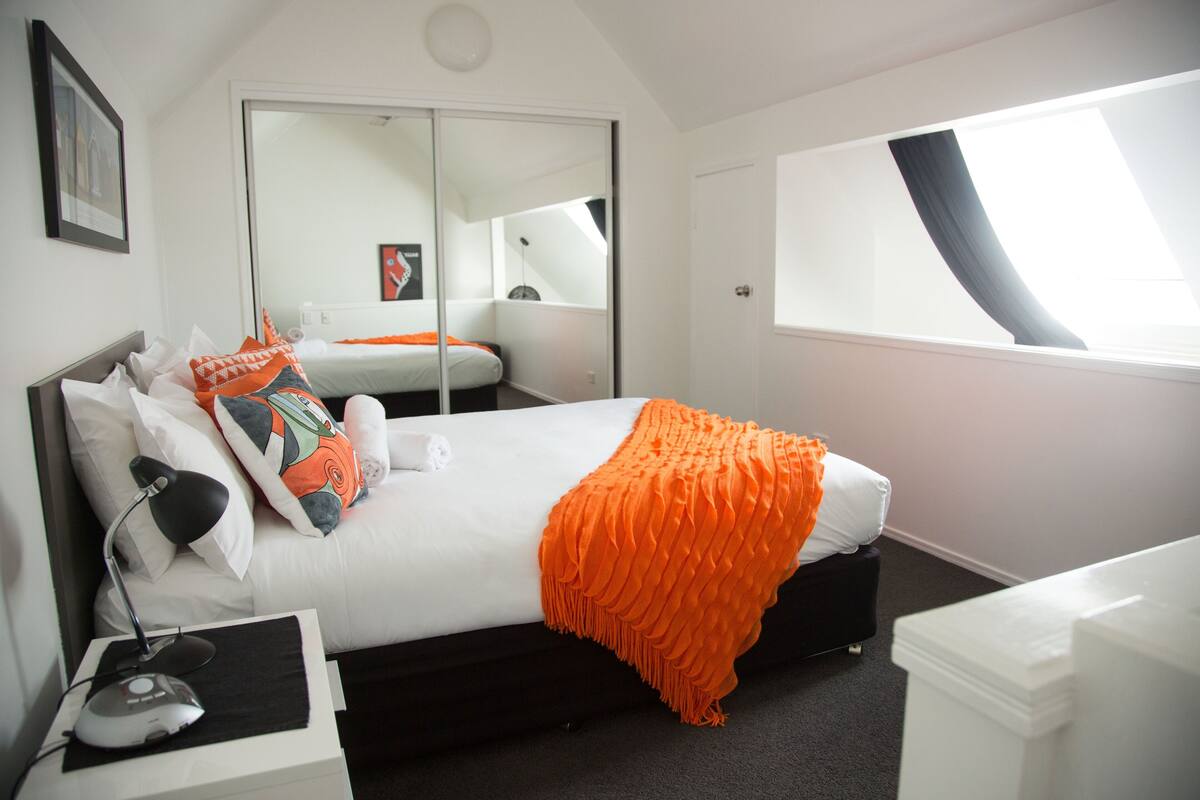 A modern hotel room with a large bed, a bedside table with a lamp, a wardrobe, and a window with a view.