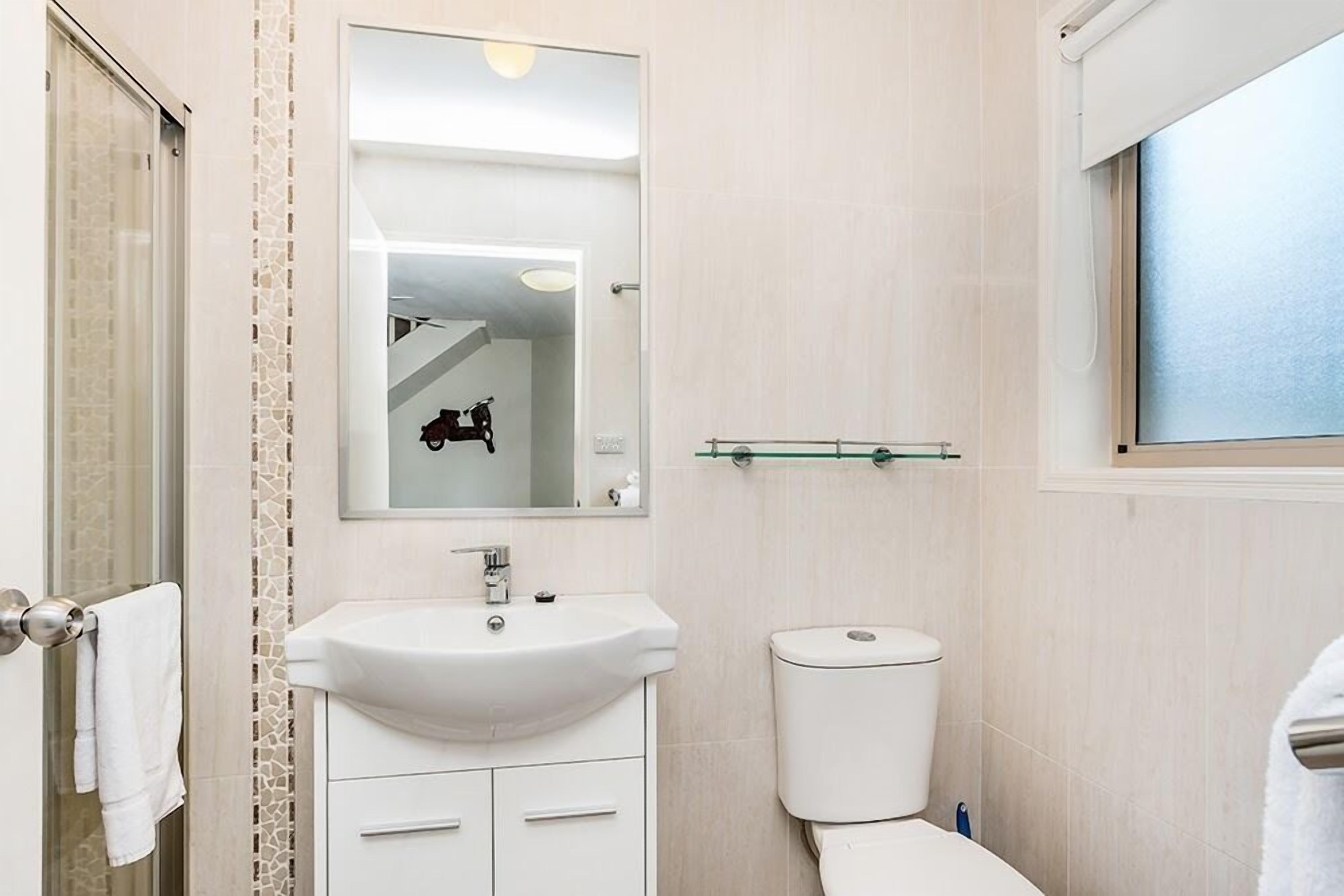 A bathroom with a white sink, mirror, and toilet, a glass shelf above the toilet, and a window with blinds.