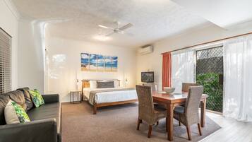 A modern hotel room with a bed, a sofa, a dining table with chairs, a ceiling fan, and a large window with curtains.